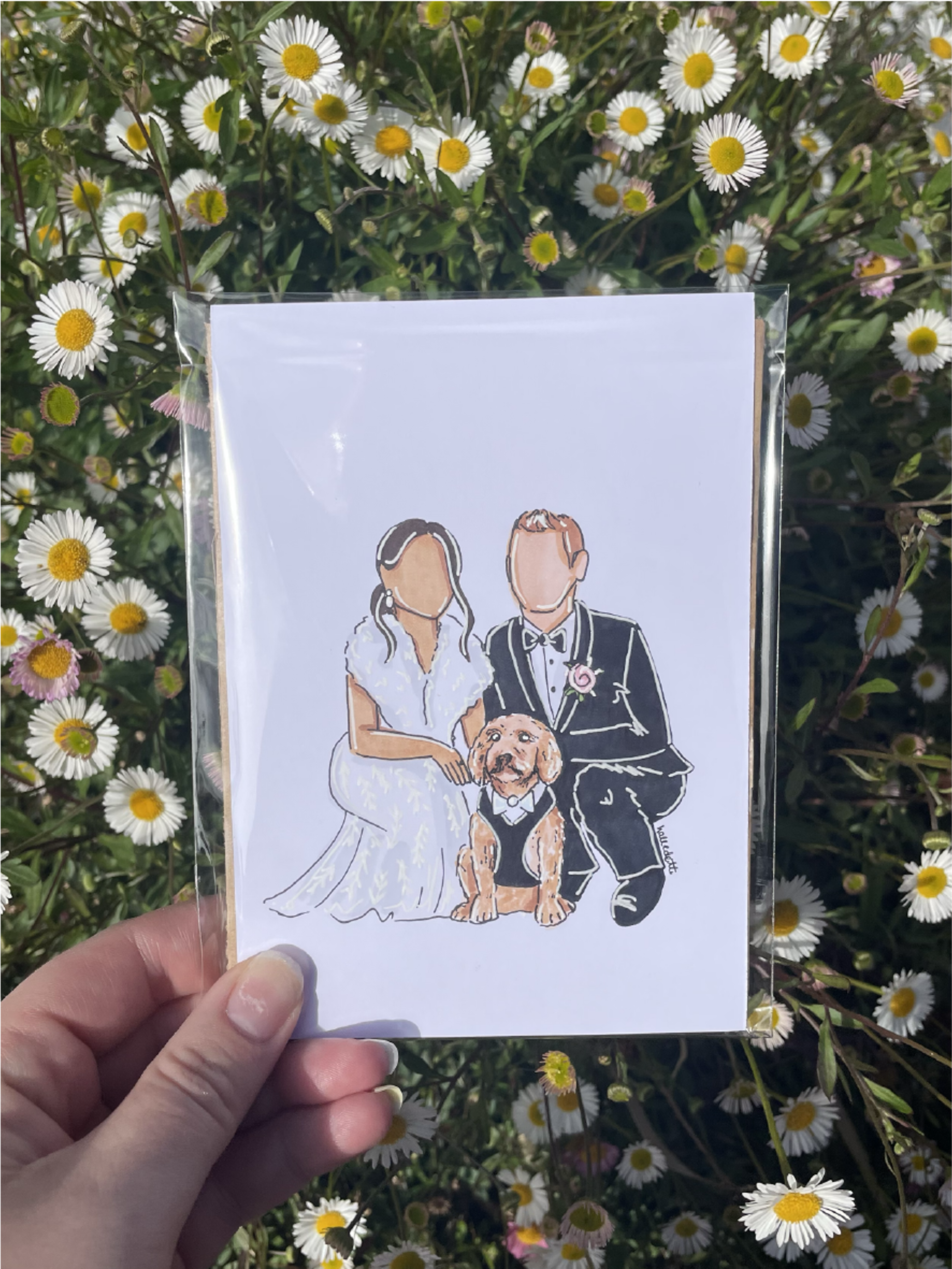 Hand holding a custom illustrated wedding portrait of a couple with their dog against a background of daisies.