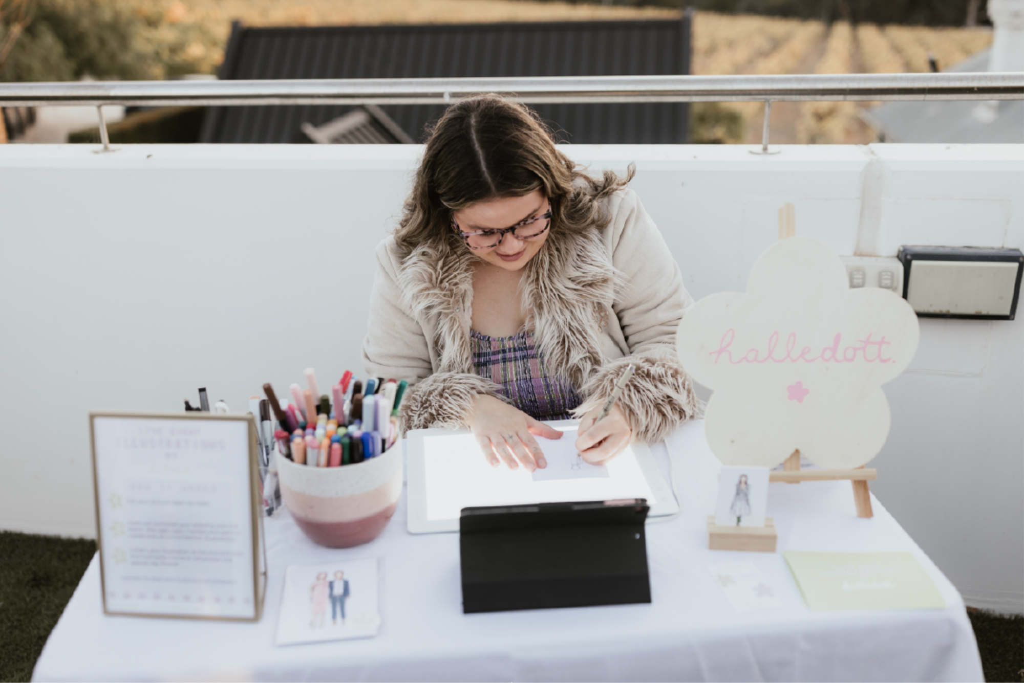 Live wedding illustrator drawing custom portraits at a white table setup with colorful markers and signage.