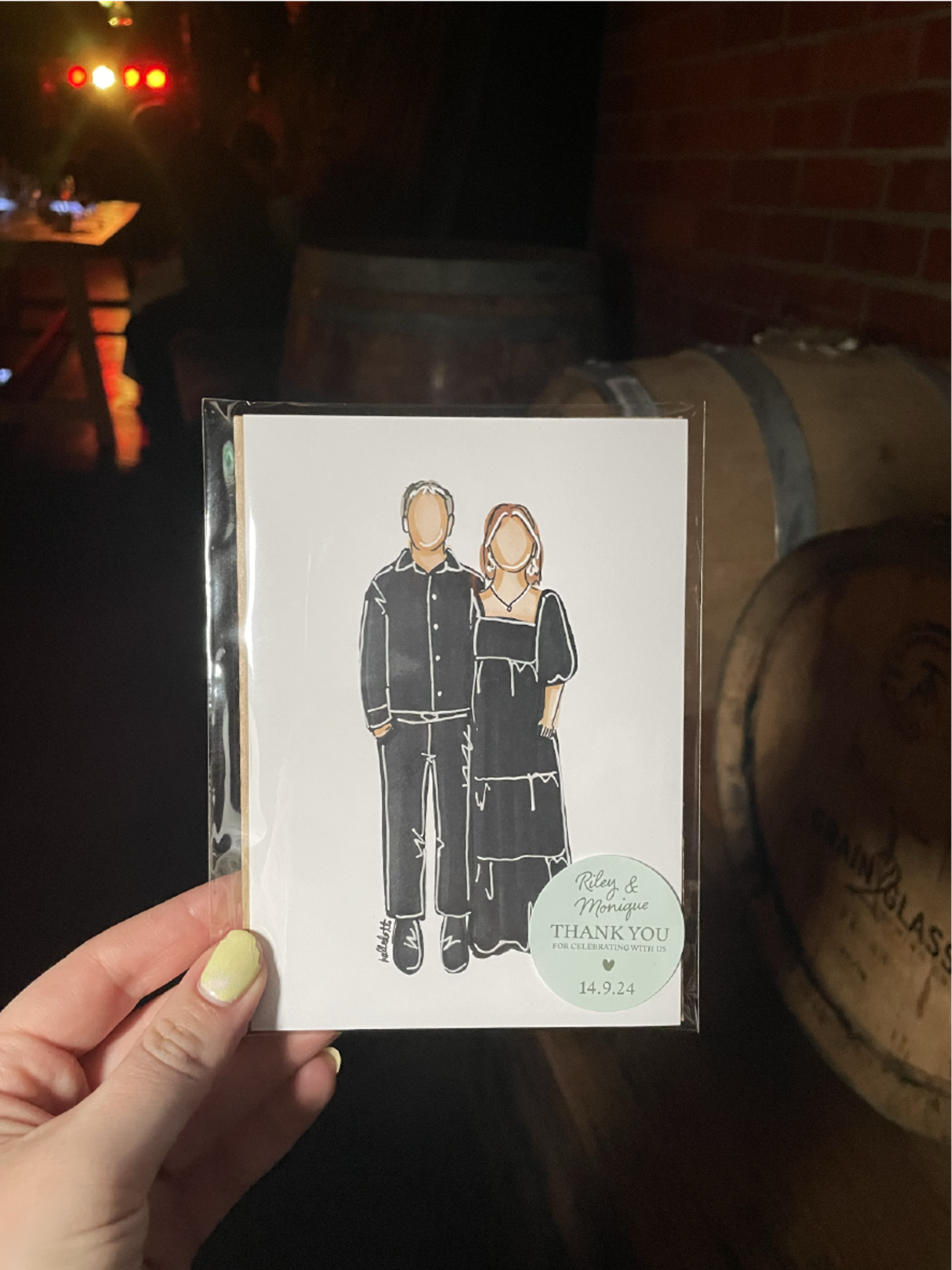 Hand holding a custom illustrated wedding thank you card of a couple in black outfits at a dimly lit venue.