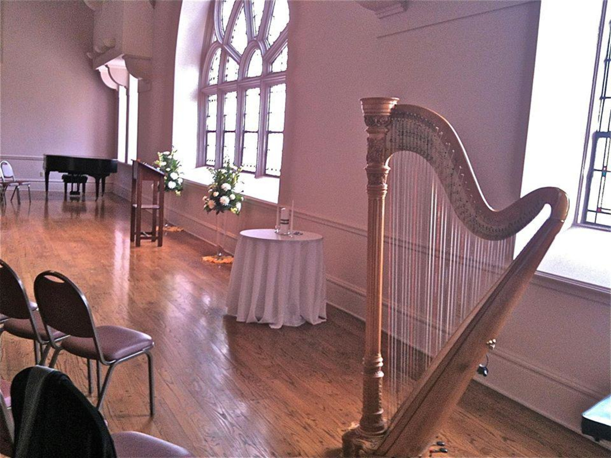 Elegant indoor wedding ceremony space with a harp, floral arrangements, and grand piano by large arched windows.