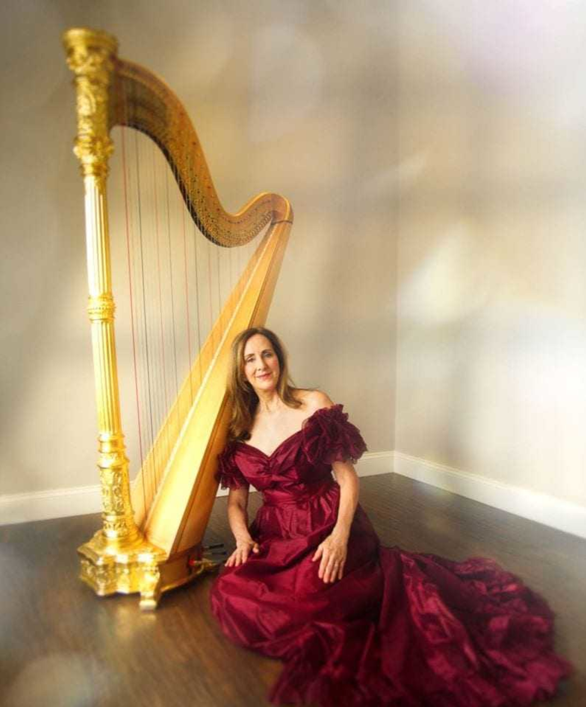 Woman in an elegant burgundy gown sits beside a tall golden harp in a softly lit room.
