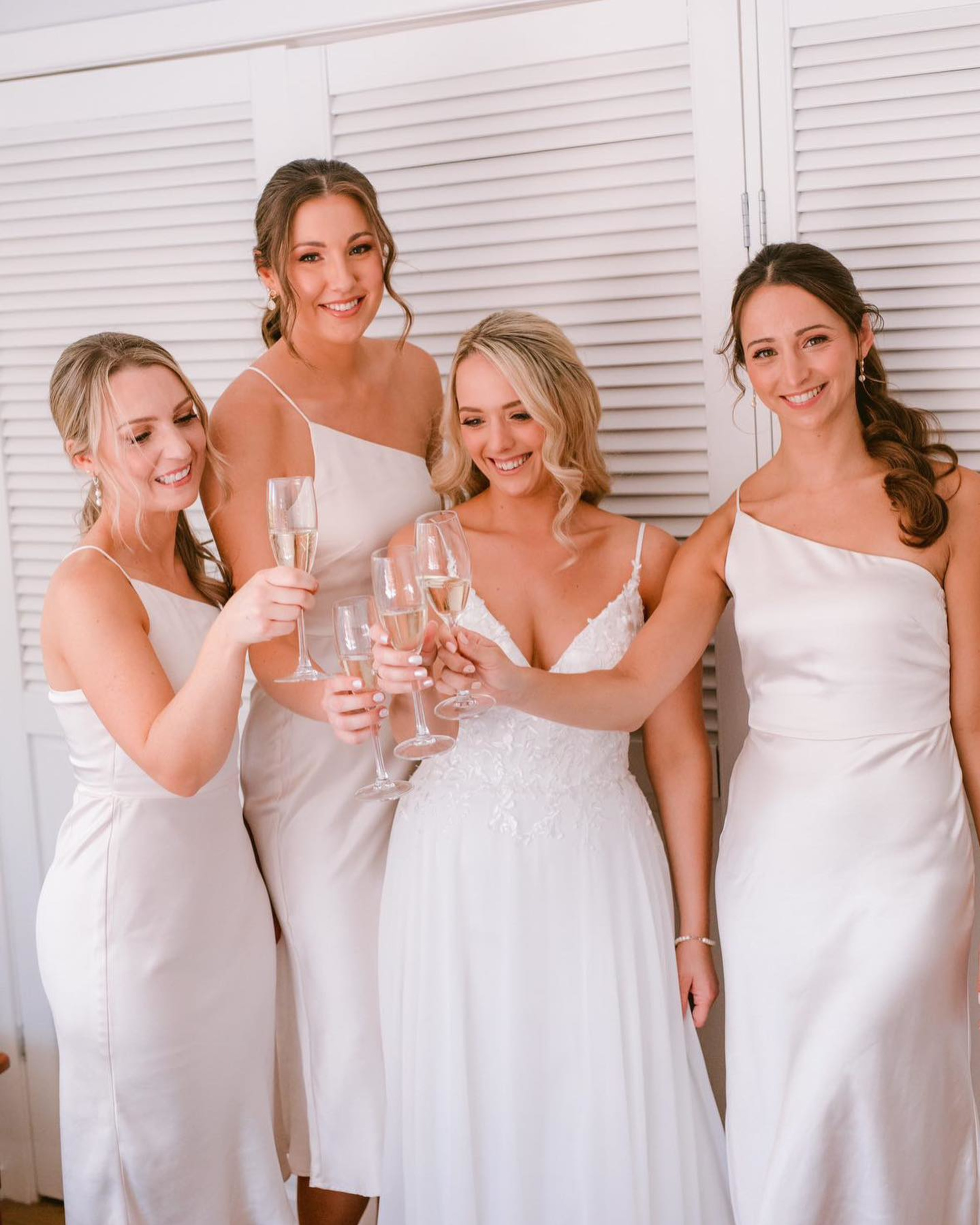 Bride and bridesmaids in ivory dresses smiling and toasting with champagne glasses indoors.