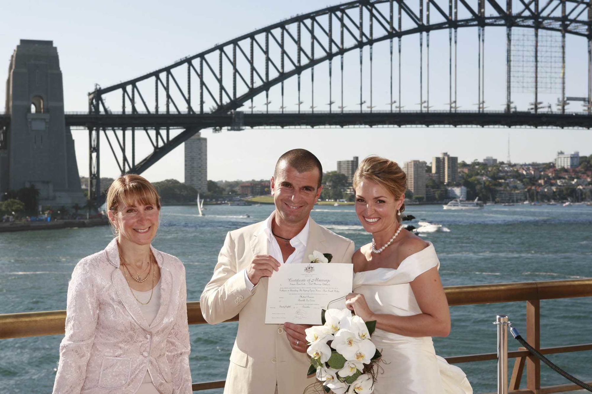 Newlywed couple with their celebrant holding a marriage certificate on a waterfront deck beneath a large bridge.