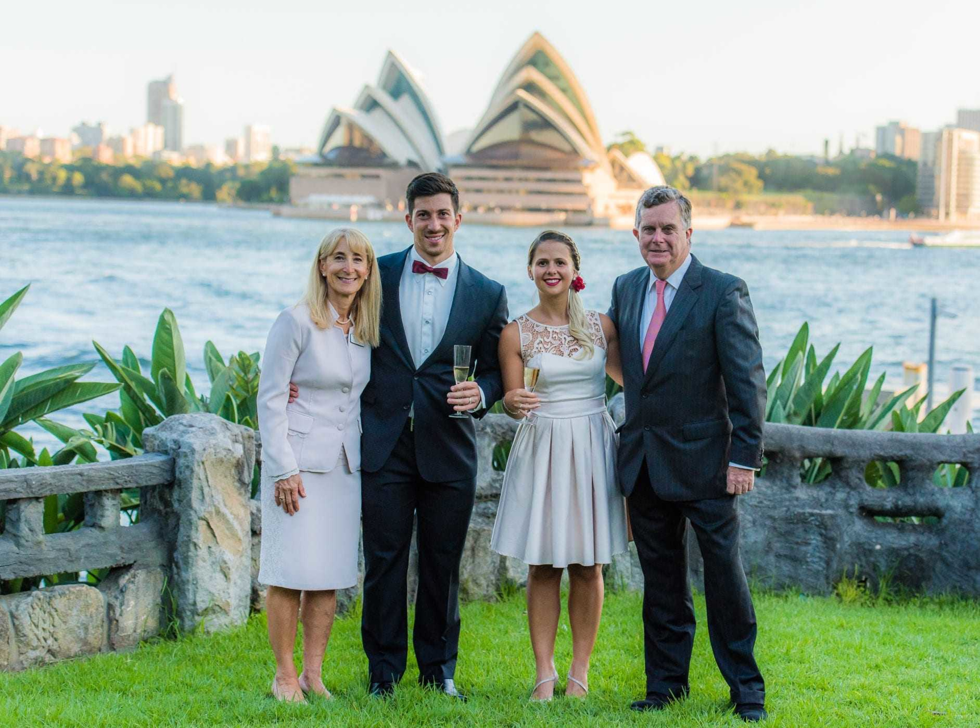 Wedding couple and parents pose for a formal portrait by the waterfront with an iconic opera house in the background.