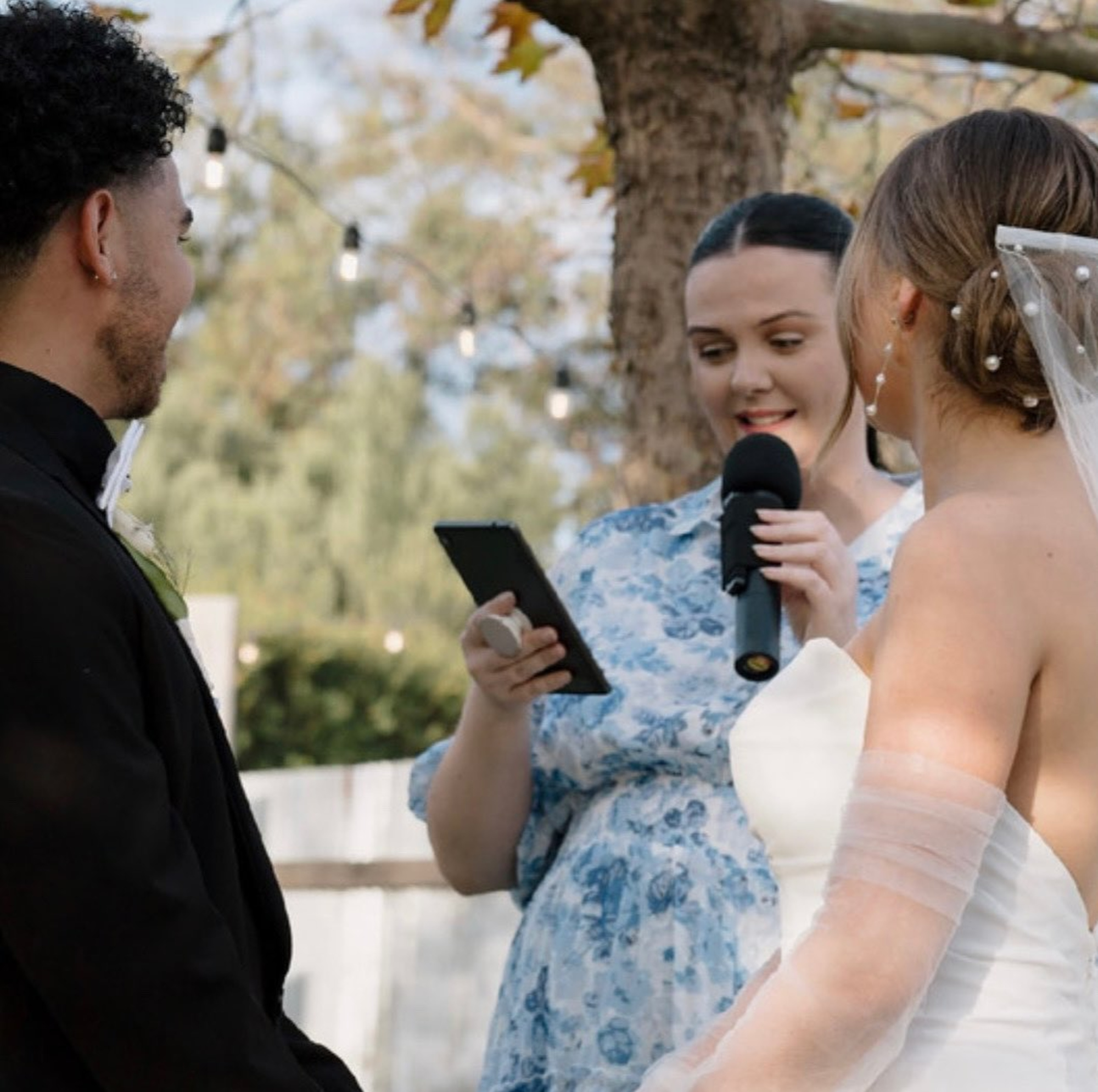 An officiant reads vows into a microphone as a bride and groom stand together during an outdoor wedding ceremony.