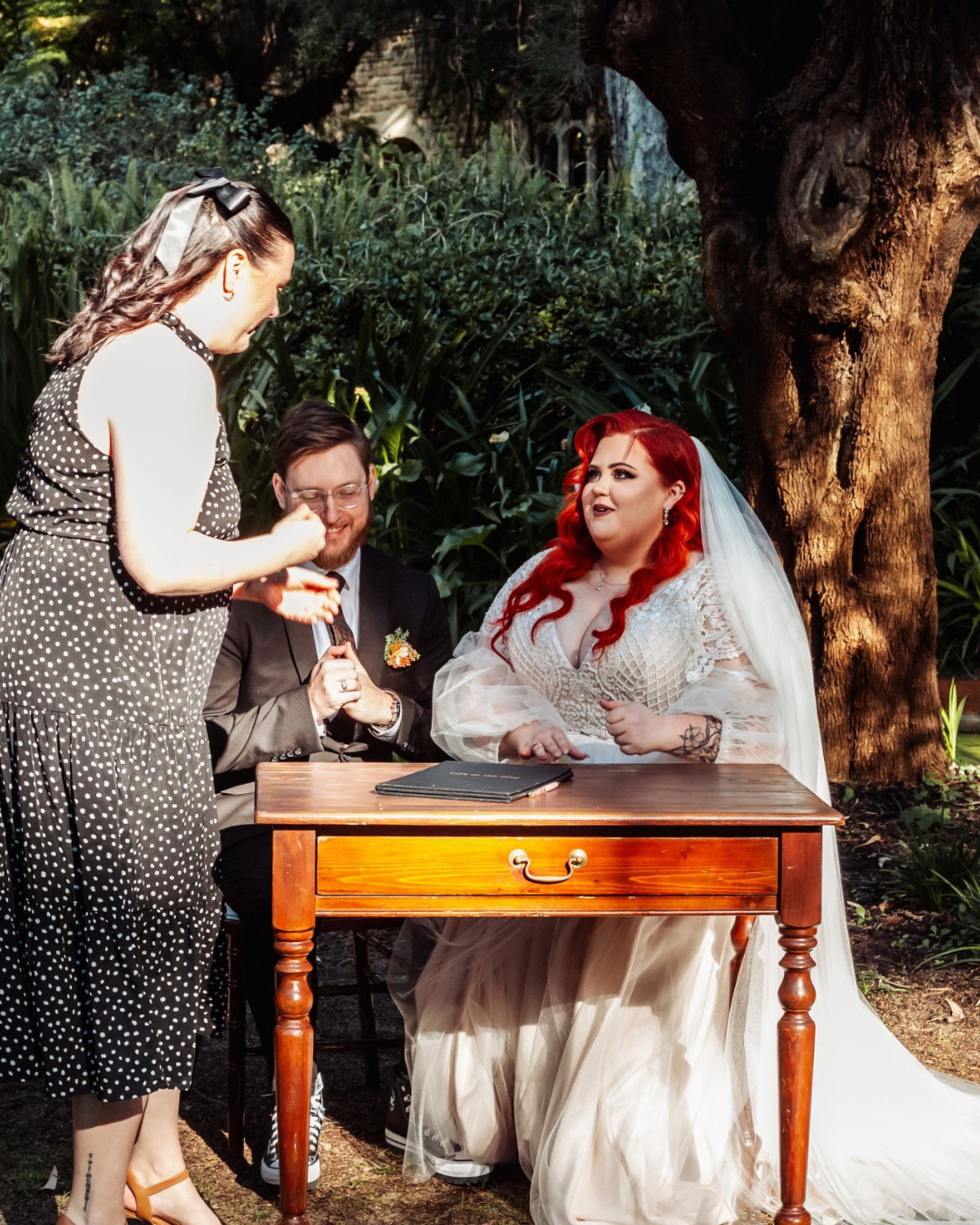 Bride and groom sit at a wooden table in a garden as an officiant guides them through signing their marriage documents.
