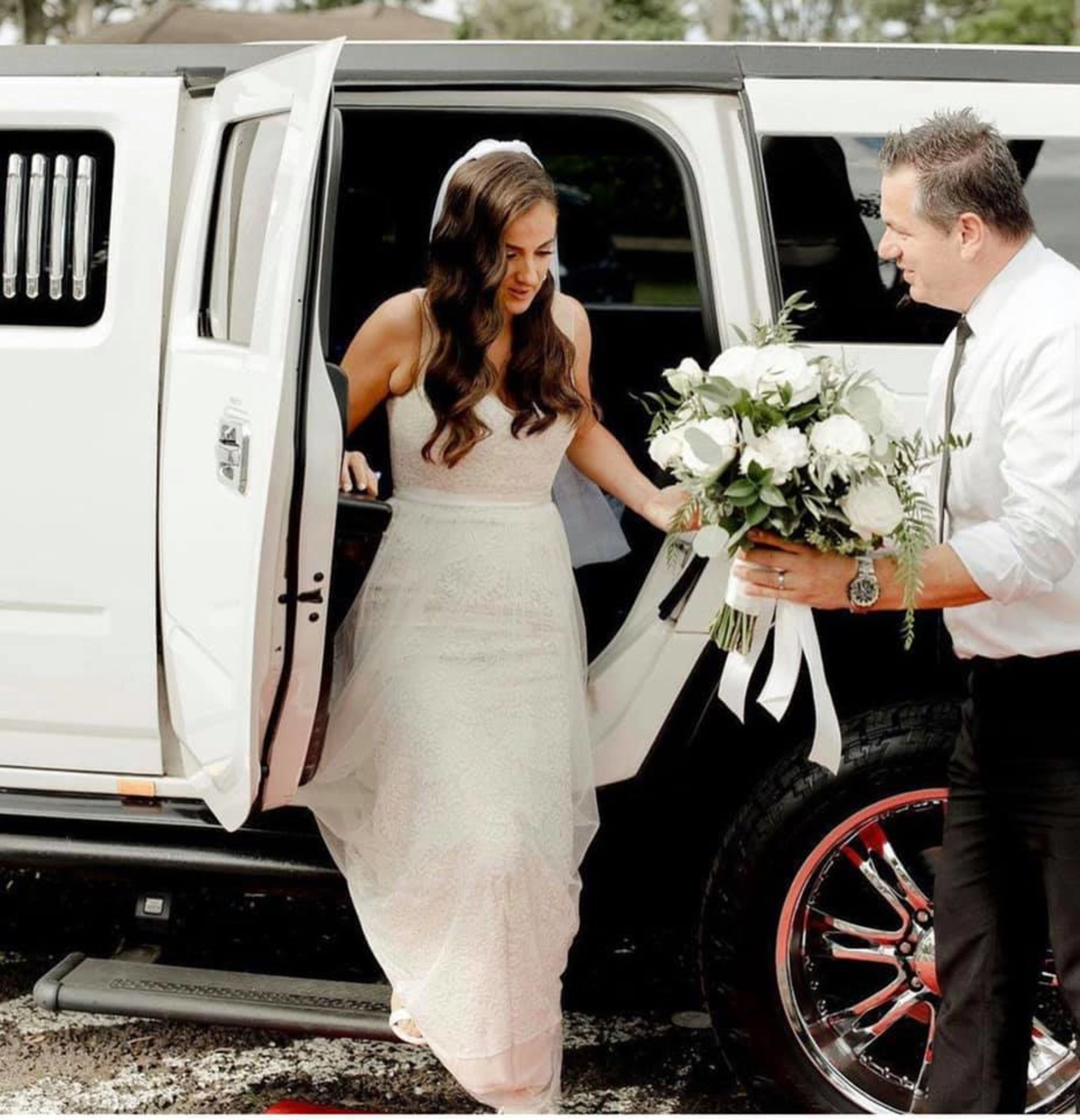 Bride stepping out of a white limousine while being handed a lush white and green bouquet.