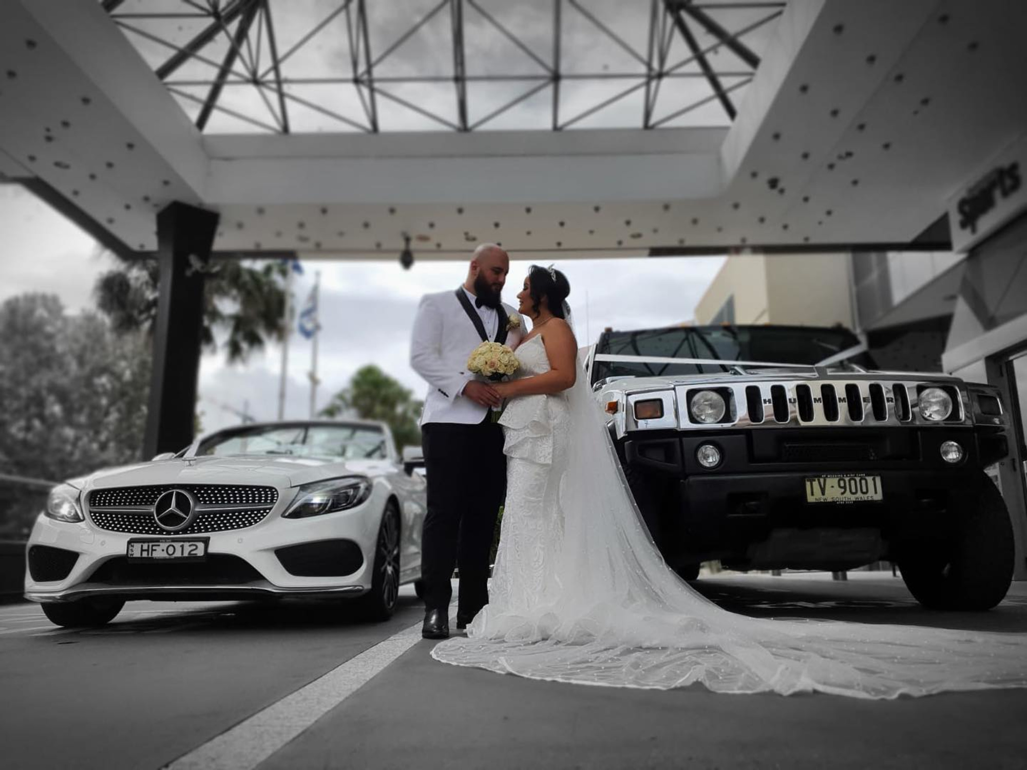 Bride and groom stand between two luxury cars outside a modern venue on their wedding day.