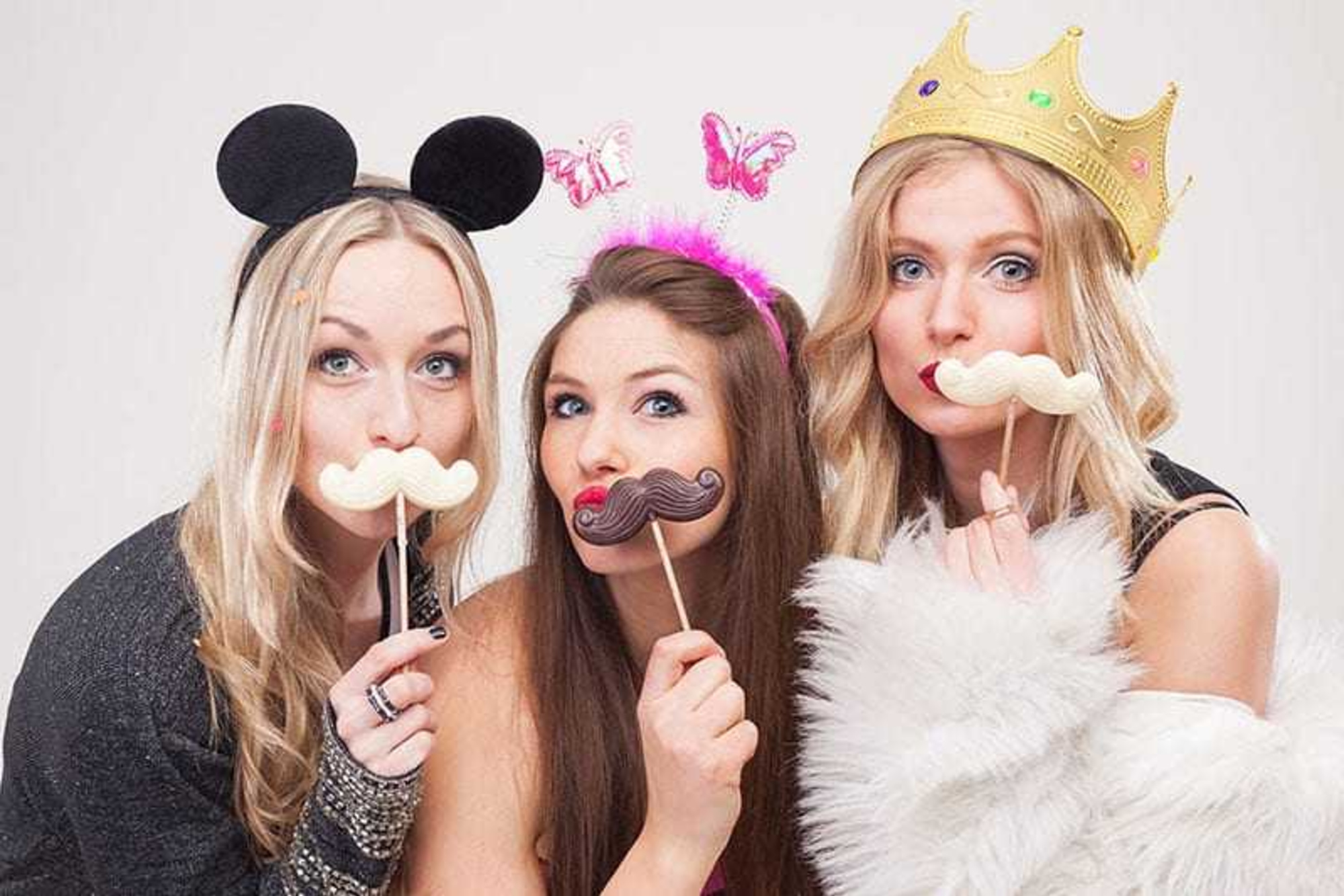 Three women posing with playful mustache props and costume headpieces in a photo booth style setup.