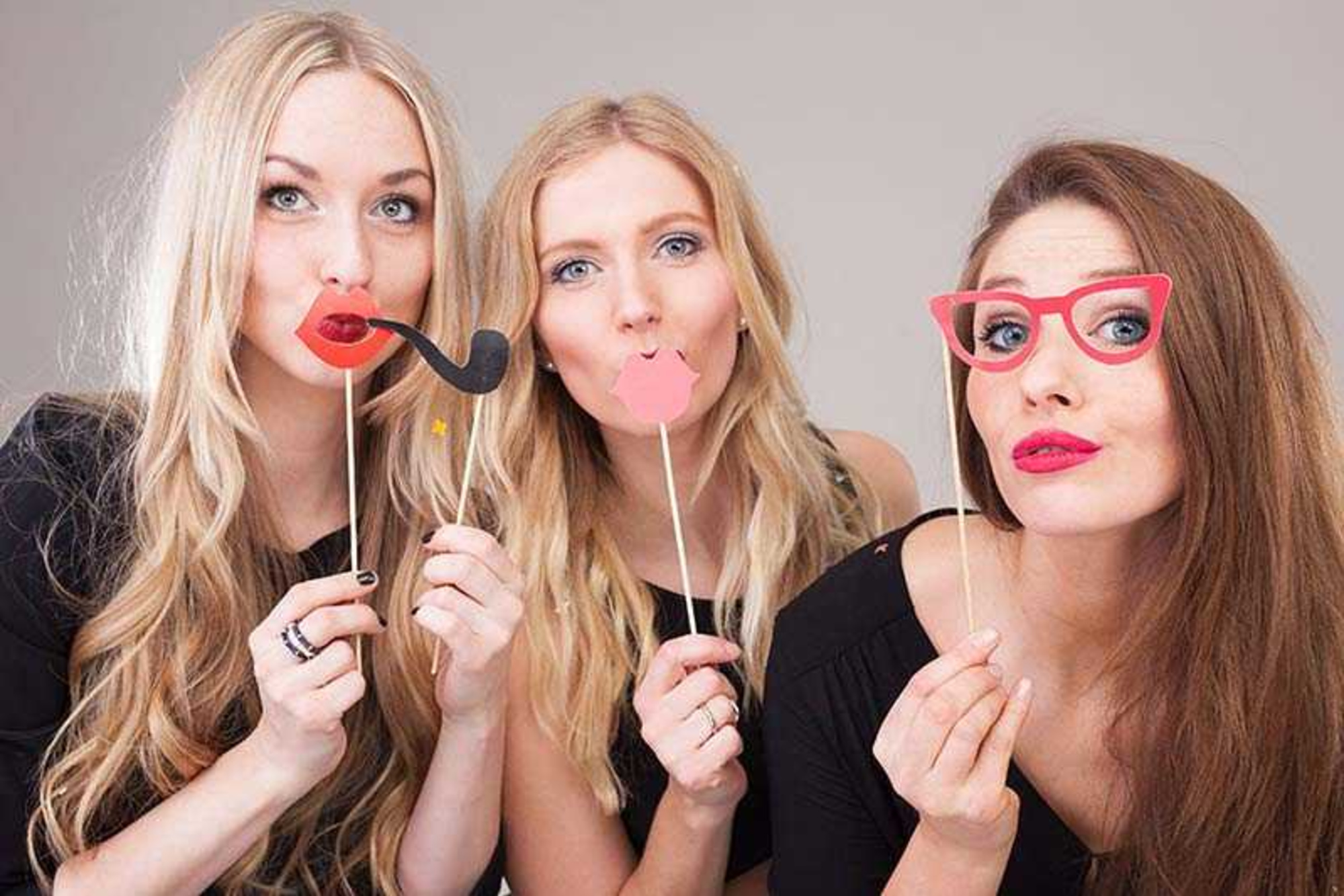 Three women pose with playful paper photo booth props in front of a neutral backdrop.