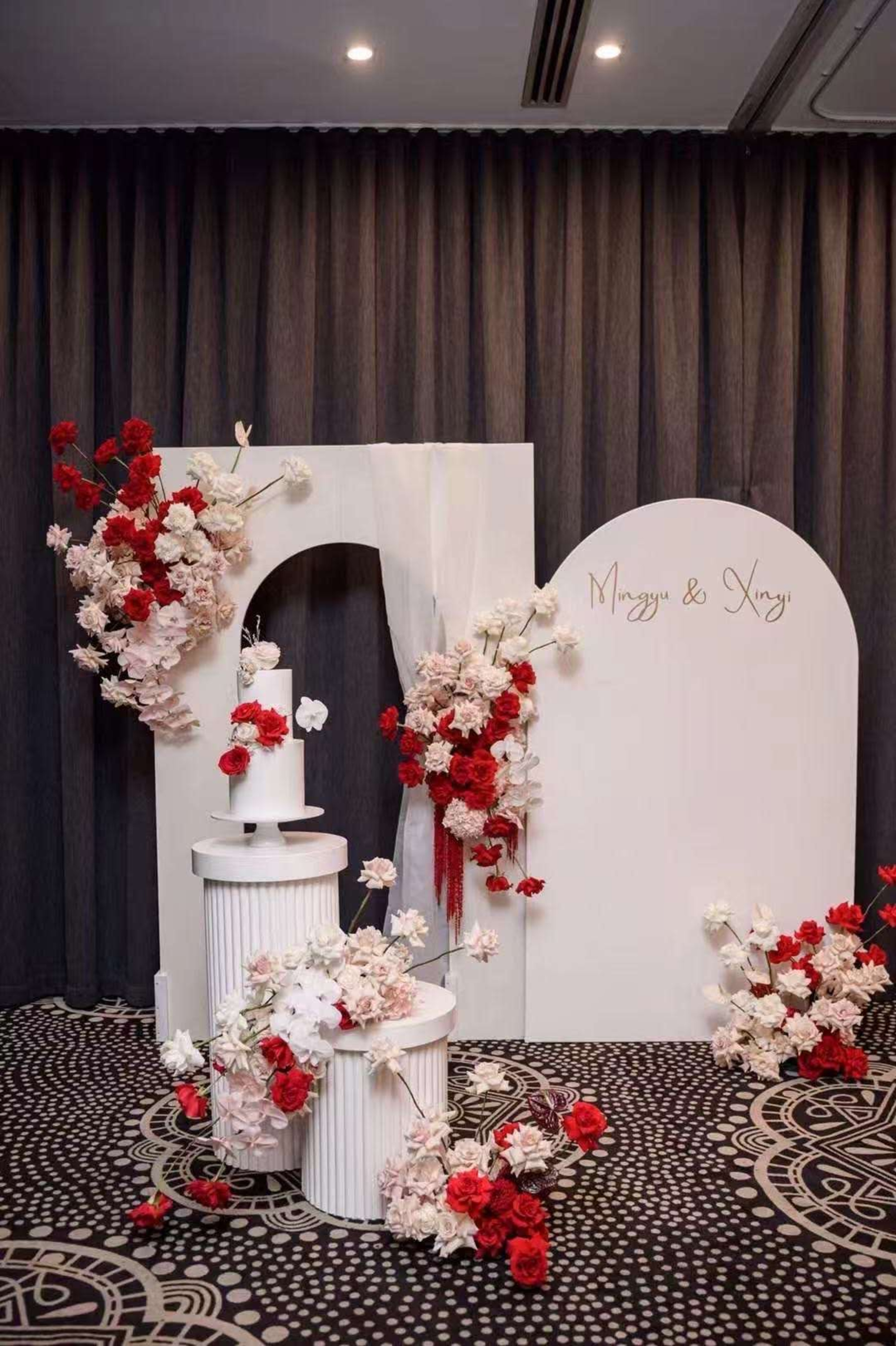 Modern indoor wedding backdrop with red and white floral arrangements and a white cake on pedestal stands.