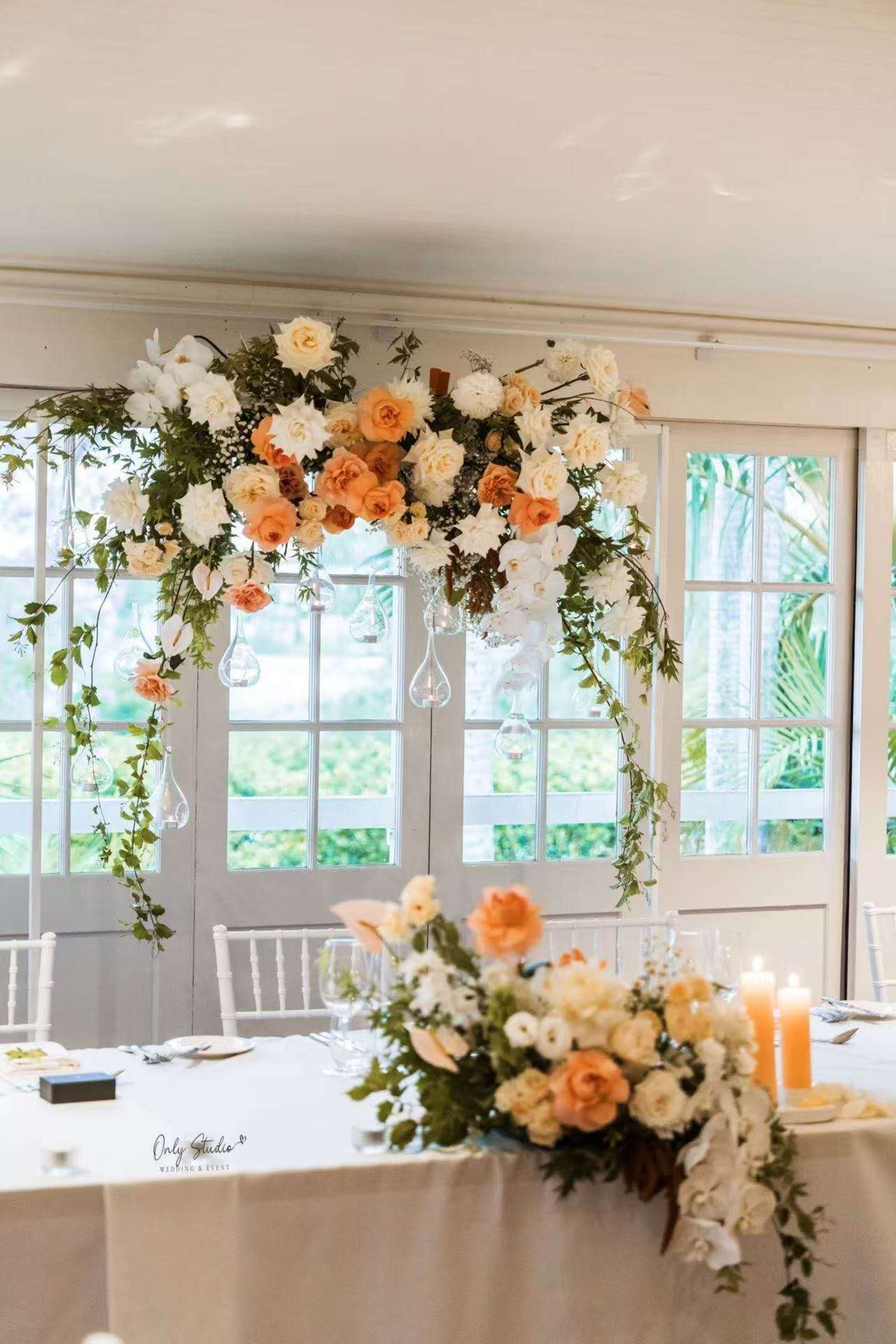 Elegant indoor wedding head table with peach and white floral arrangements and hanging greenery in front of tall windows.