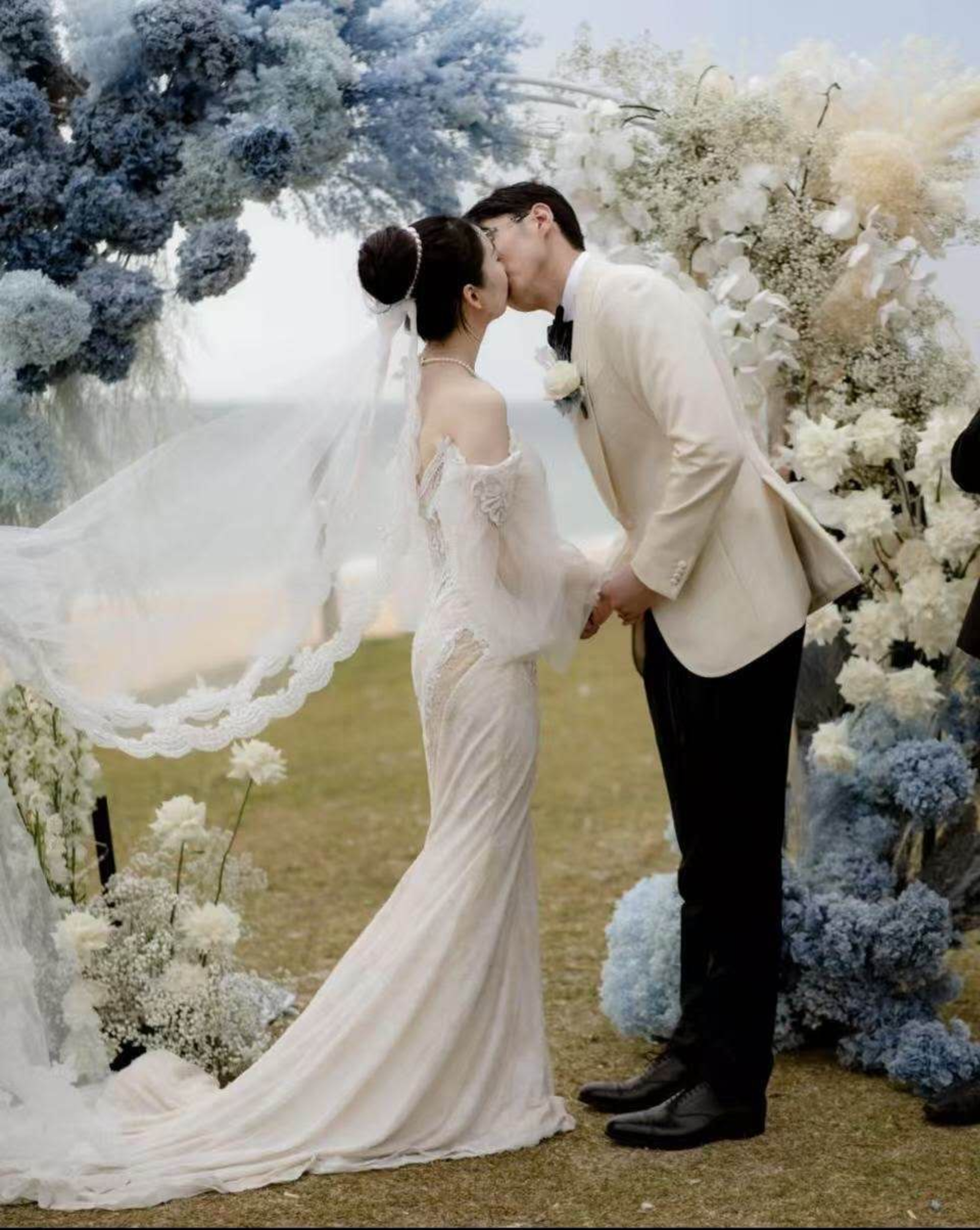Bride and groom share a kiss beneath a blue and white floral wedding arch outdoors.