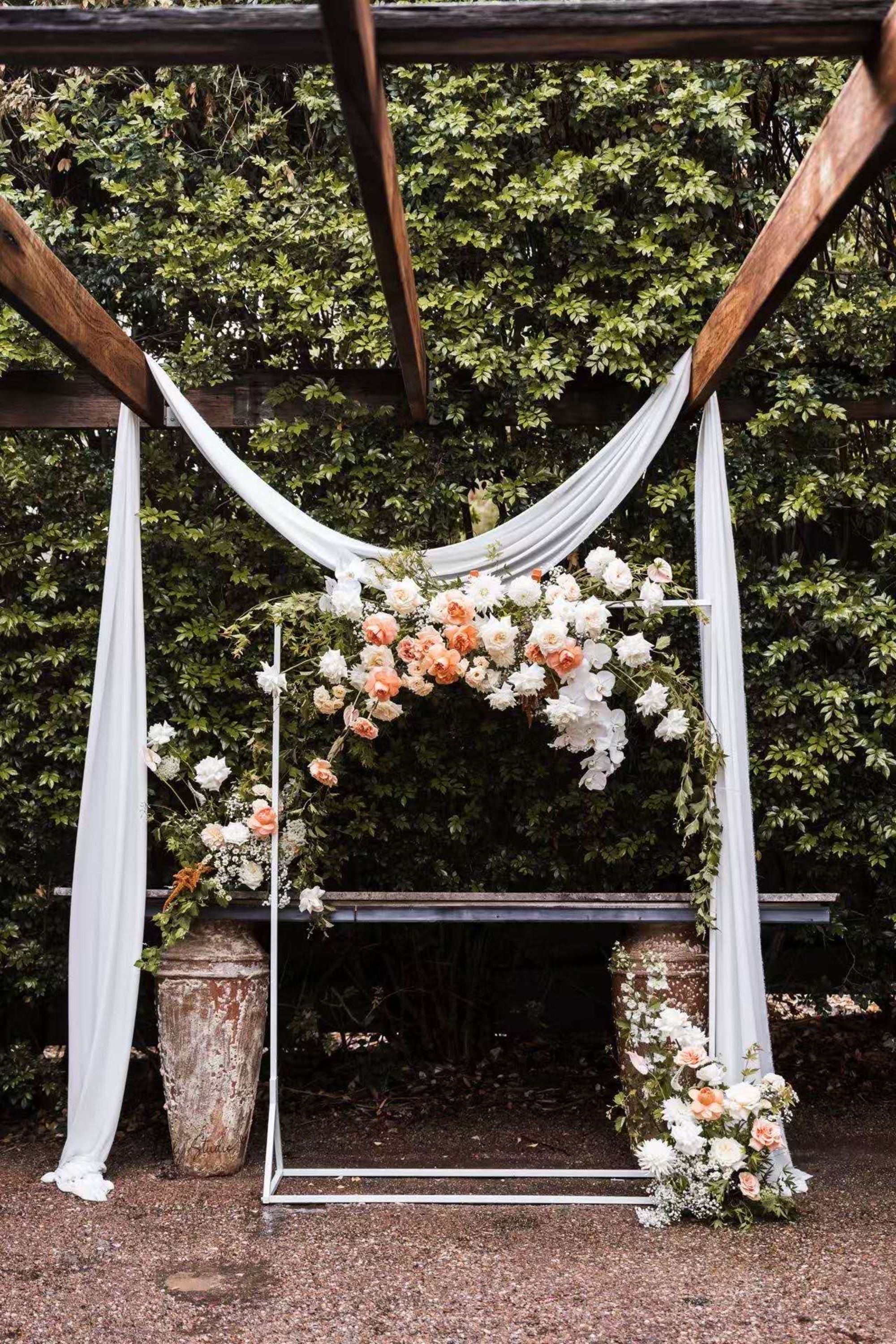 Outdoor wedding ceremony arch with white draping and peach and white floral arrangements under wooden beams.
