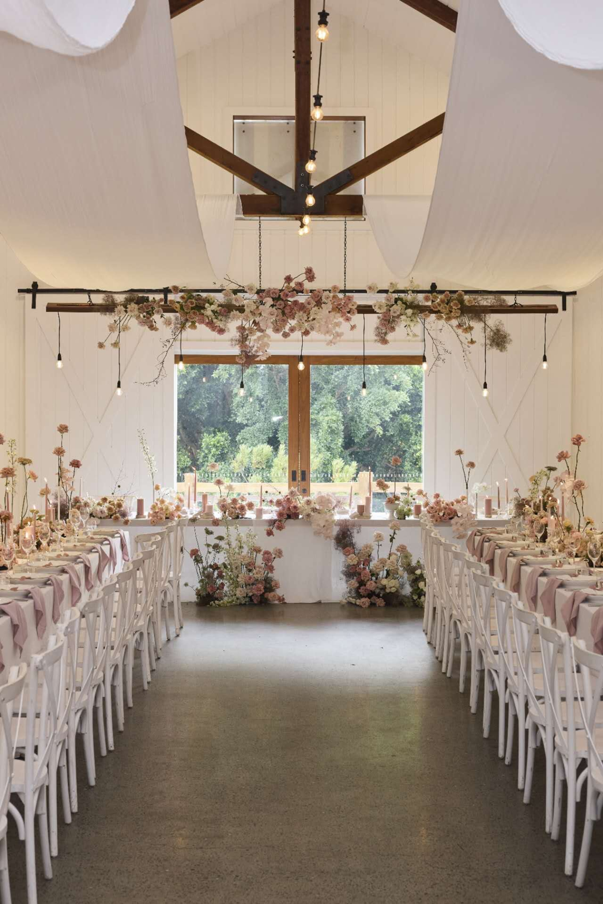 Romantic indoor wedding reception with long banquet tables, pastel florals, and hanging lights in a bright barn-style venue.