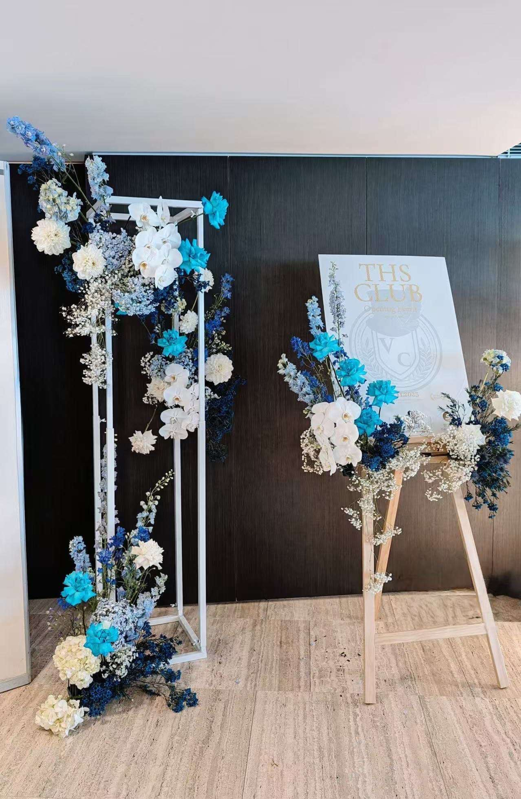 Modern indoor wedding welcome display with blue and white floral arrangements around a sign and geometric stand.