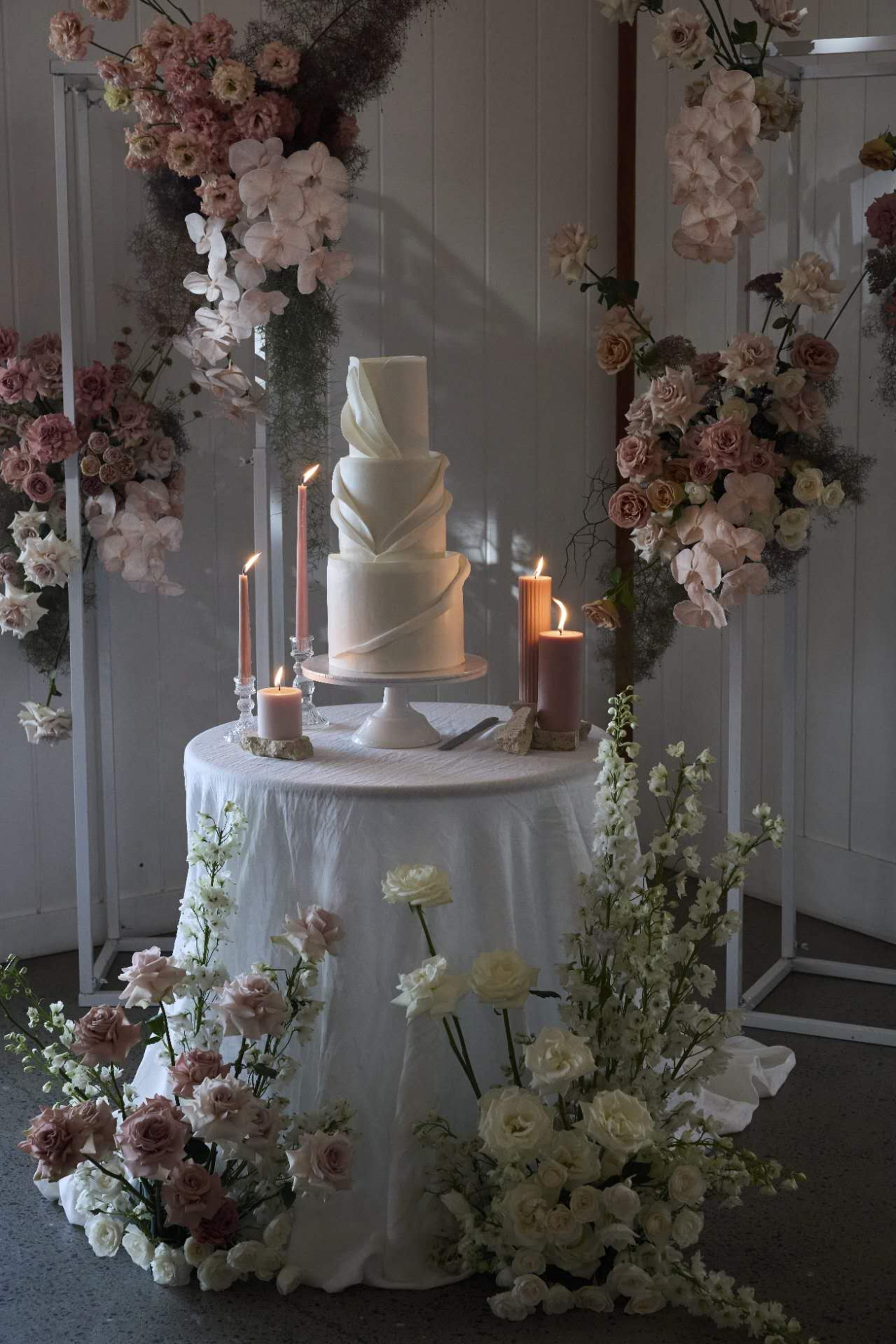 Tall white wedding cake on a round table surrounded by pastel florals and glowing candles in a romantic setup.