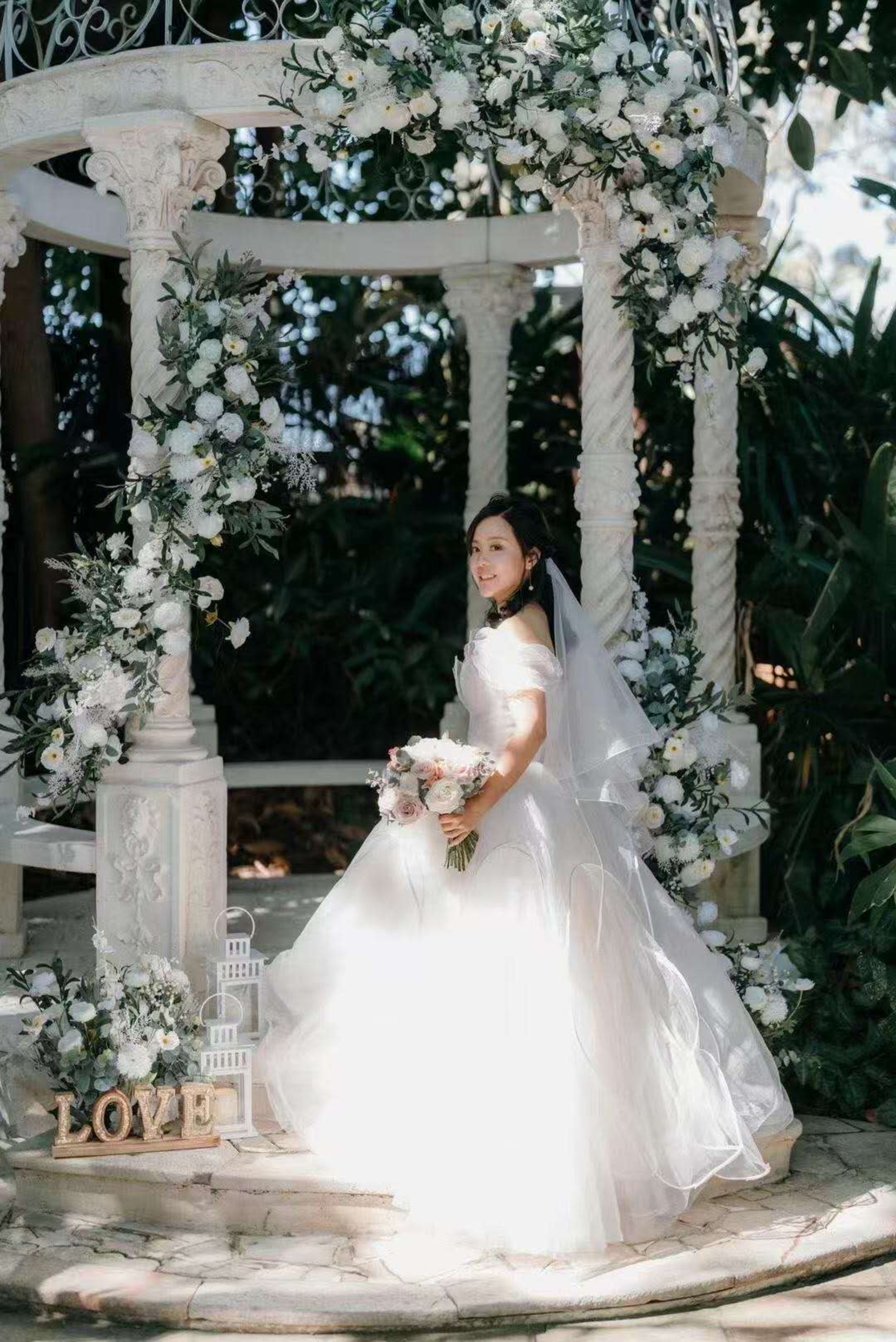 Bride in a white gown poses with bouquet under a flower-covered gazebo in a garden setting.