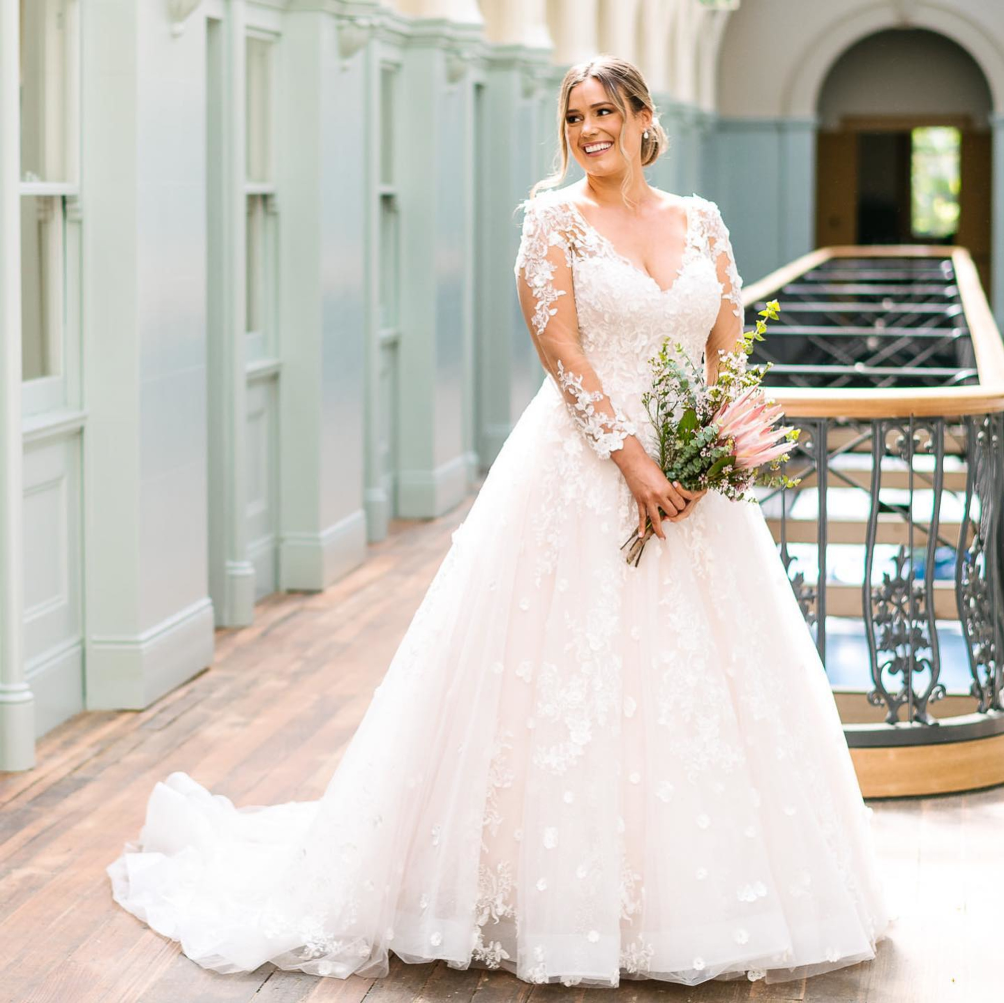 Smiling bride in a long-sleeve lace wedding gown holding a bouquet in a bright indoor venue hallway.