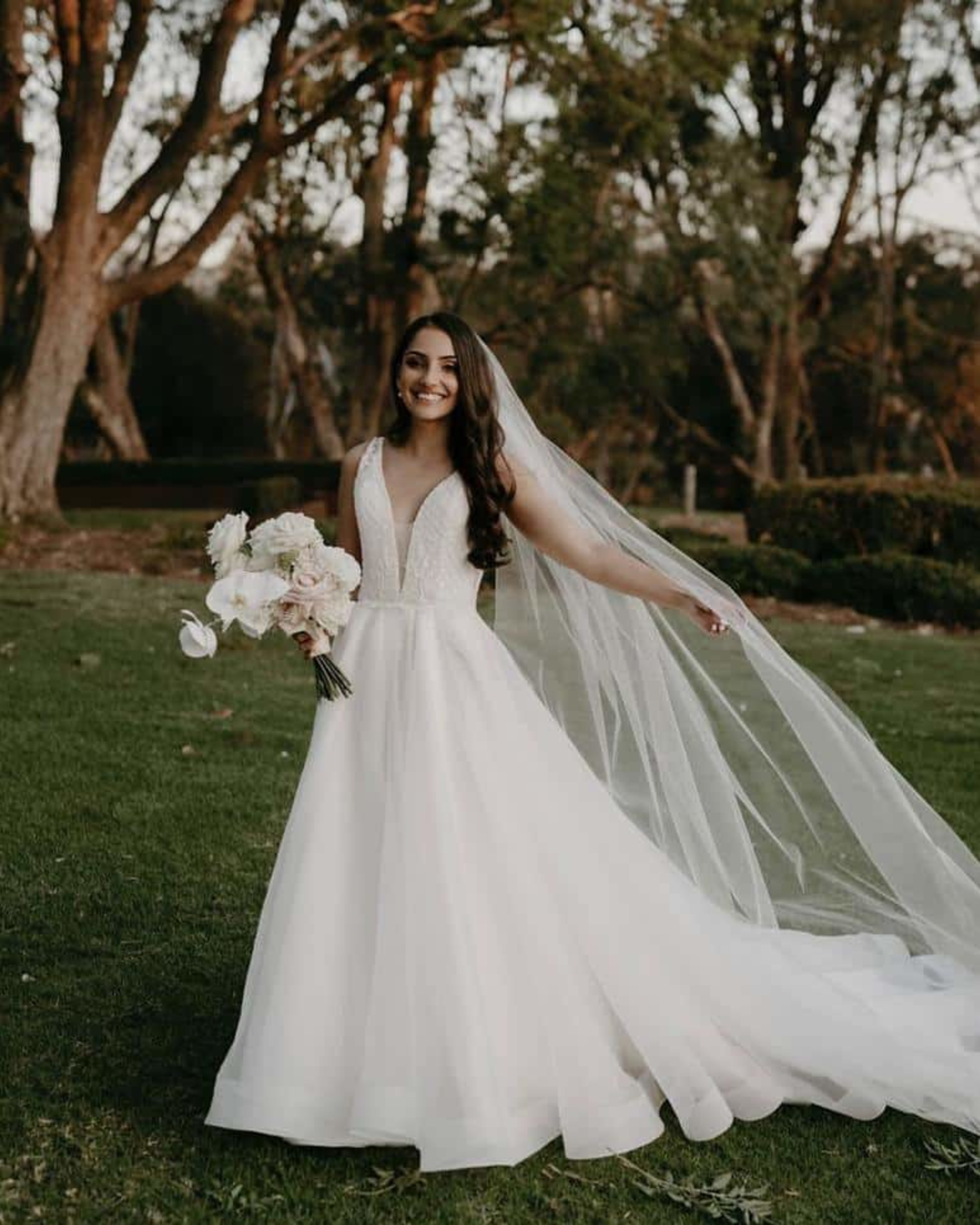 Smiling bride in a flowing white gown and long veil holding a bouquet in a garden setting.