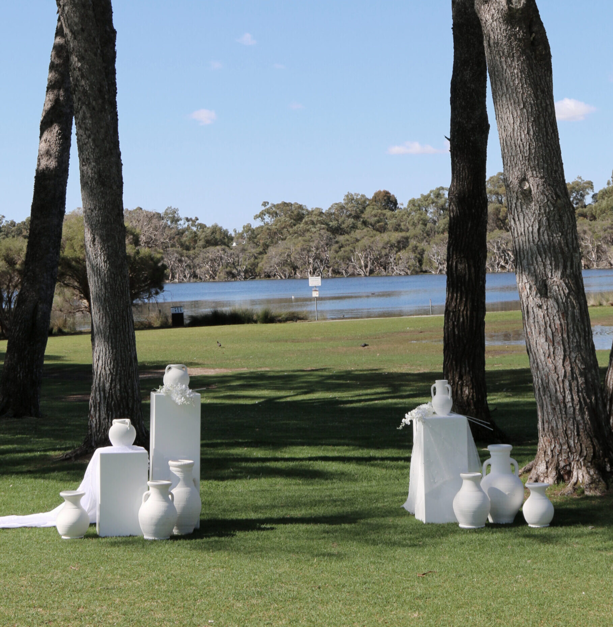 Minimalist white vase wedding ceremony setup on a grassy lakeside lawn beneath tall trees.