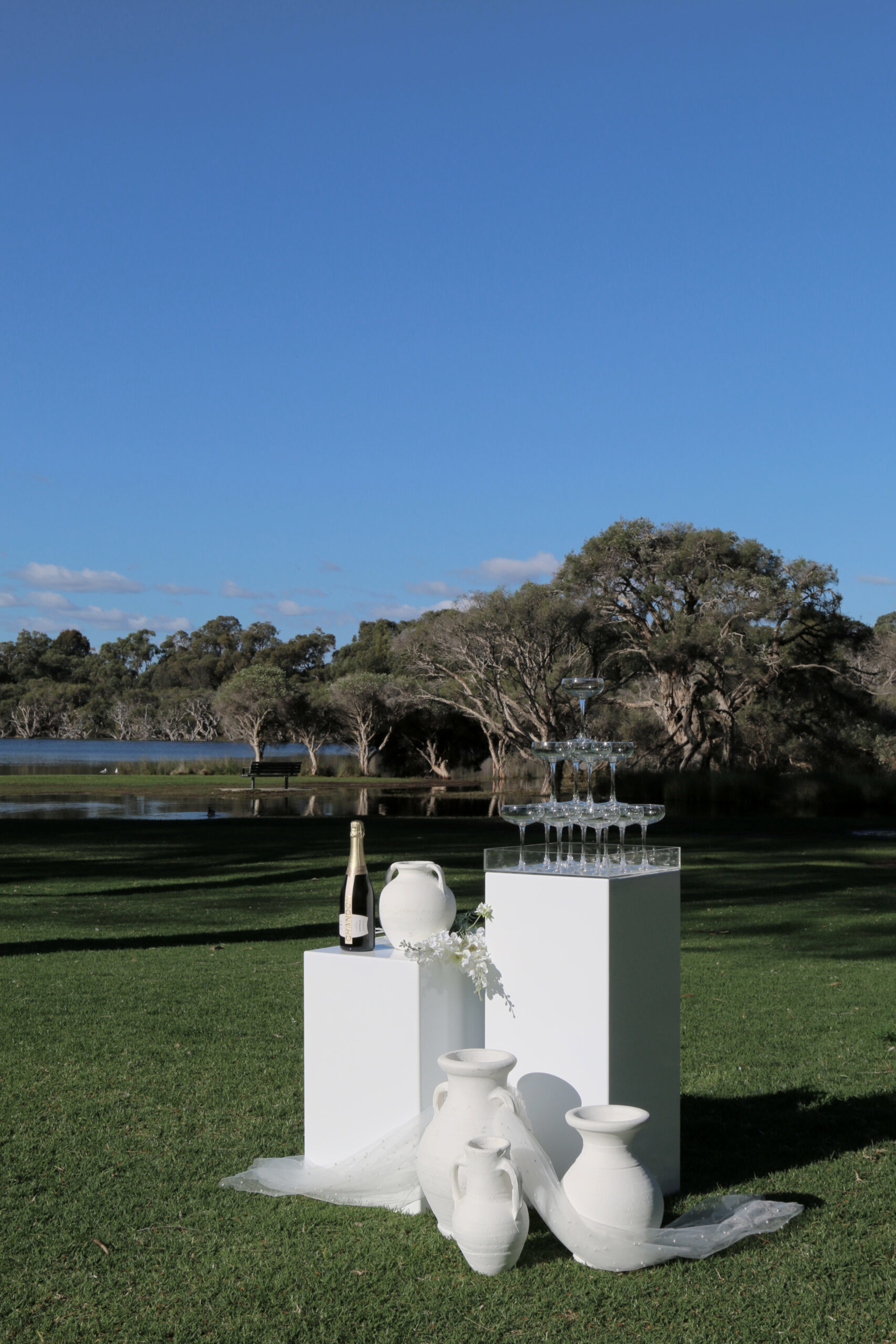 Minimalist white pedestal display with champagne tower and vases on a lawn by a lakeside backdrop.