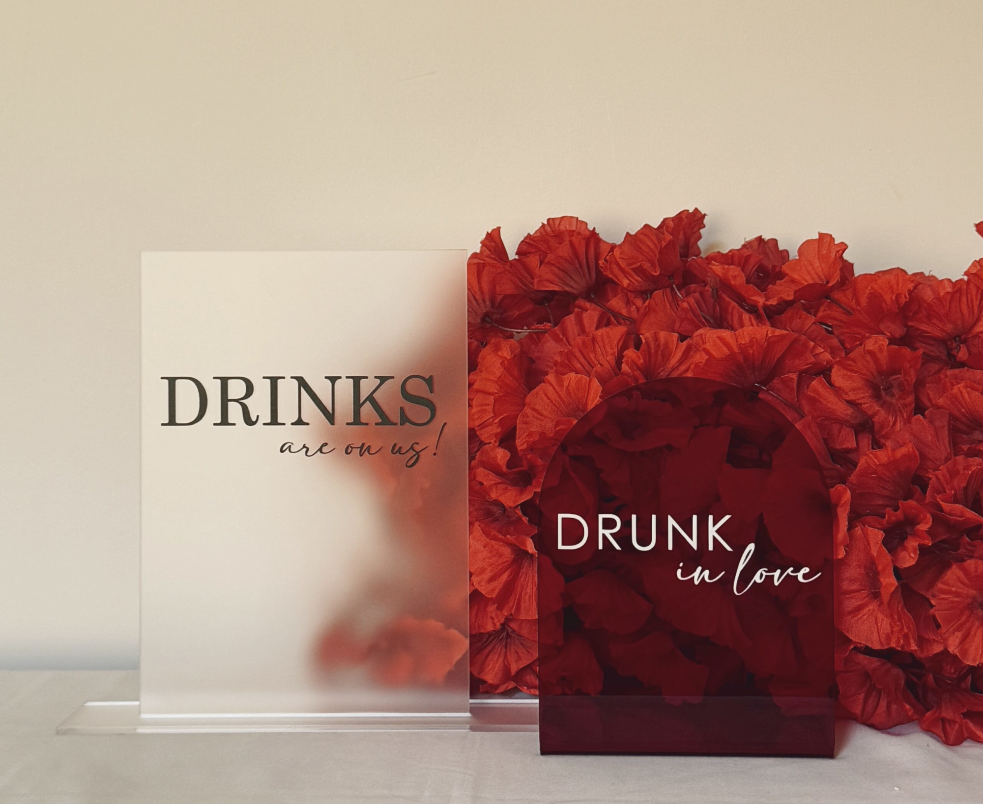 Modern acrylic wedding bar signs reading drinks are on us and drunk in love against a backdrop of red flowers.