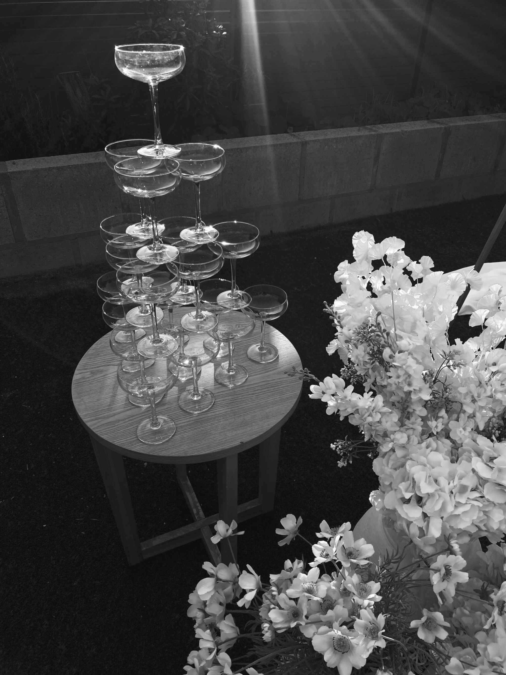Champagne coupe tower on a small wooden table beside lush flowers at an outdoor wedding.