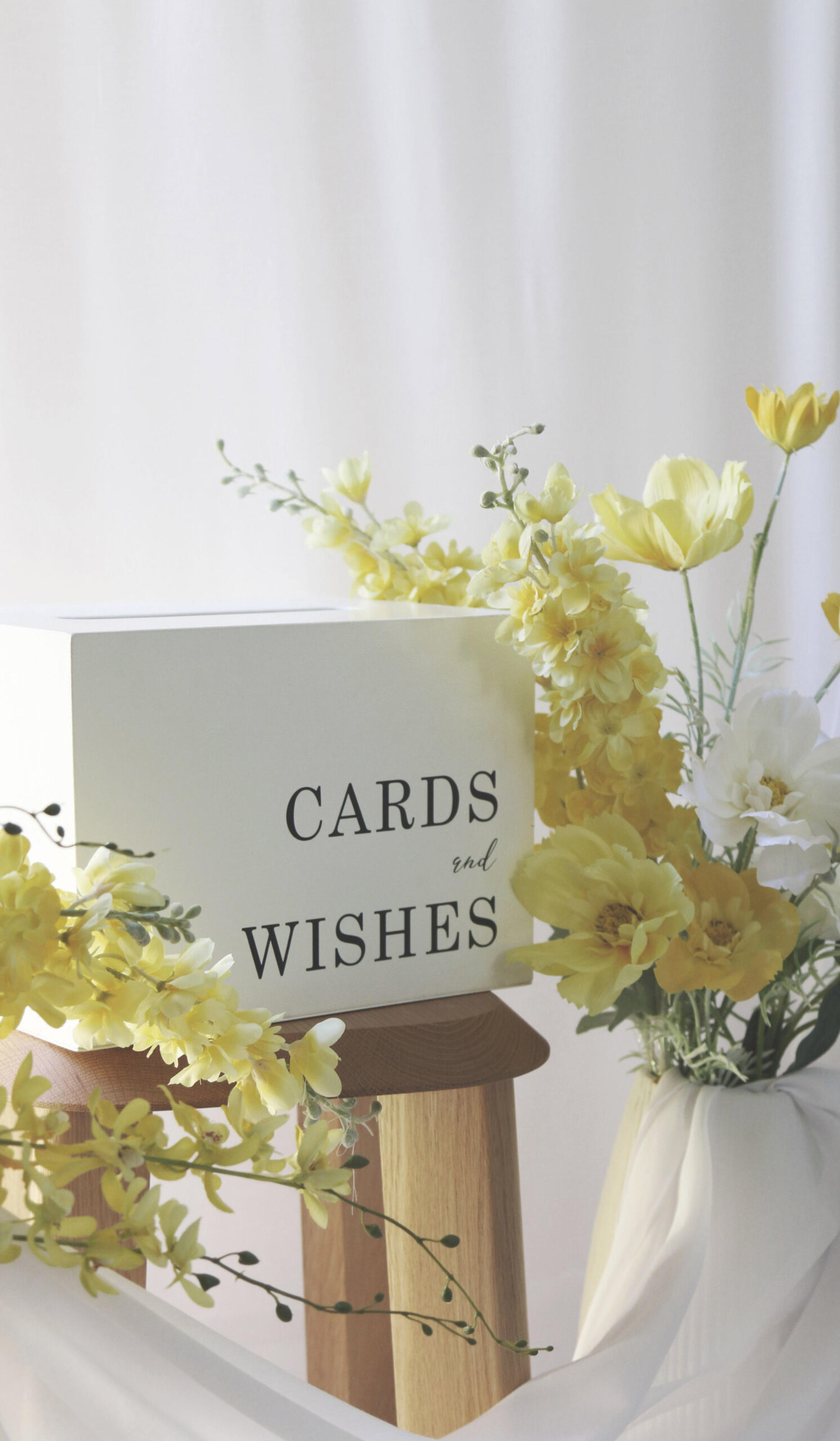 Wedding cards and wishes box styled with yellow flowers on a wooden stool against a soft white backdrop.