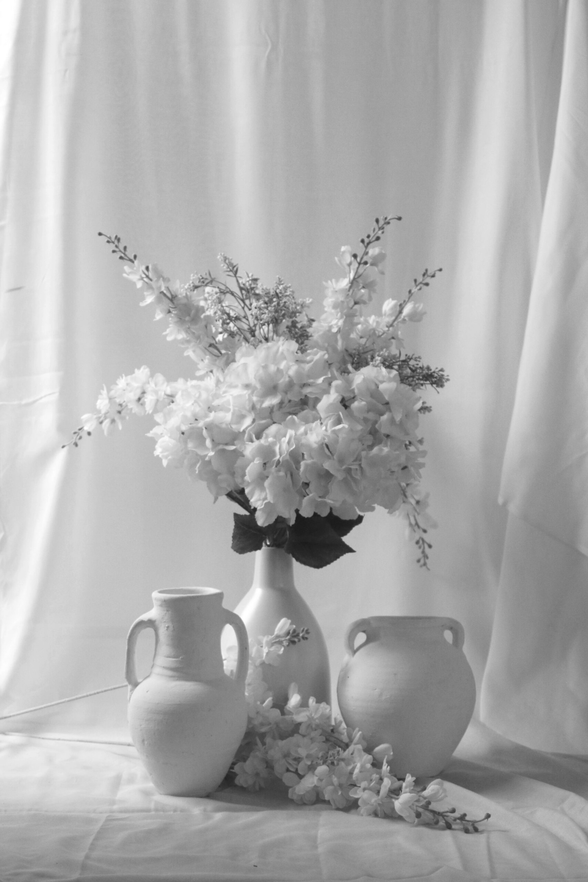 Monochrome arrangement of white flowers in a vase with two ceramic jugs on a draped fabric backdrop.