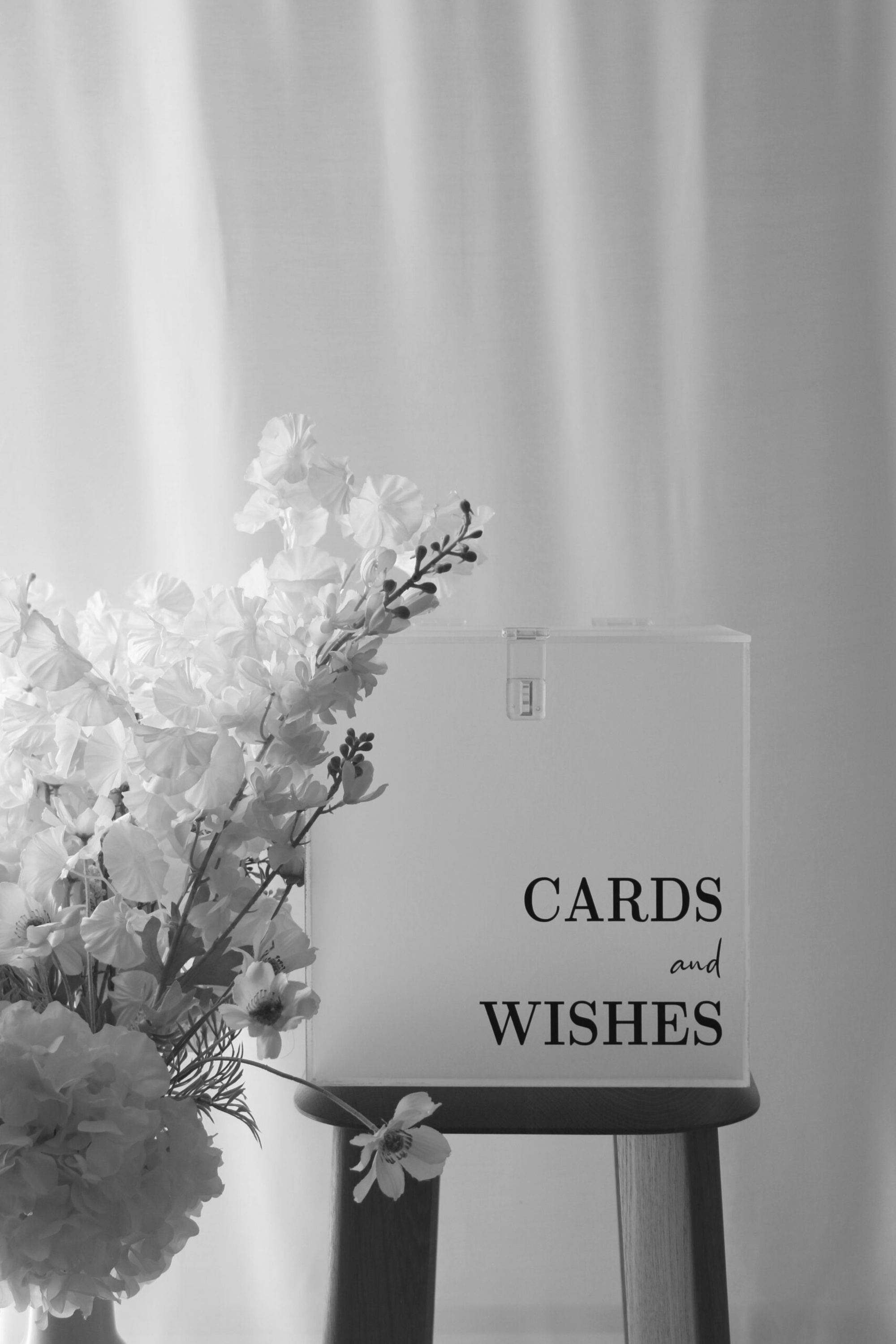 Wedding cards and wishes box on a wooden stand beside white floral arrangements in soft light.