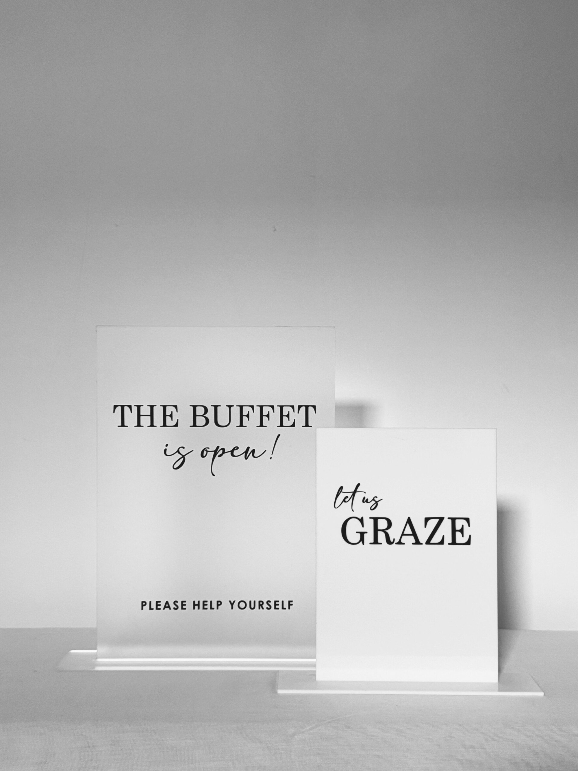 Minimalist acrylic buffet and grazing table signs for a wedding reception displayed on a neutral surface.