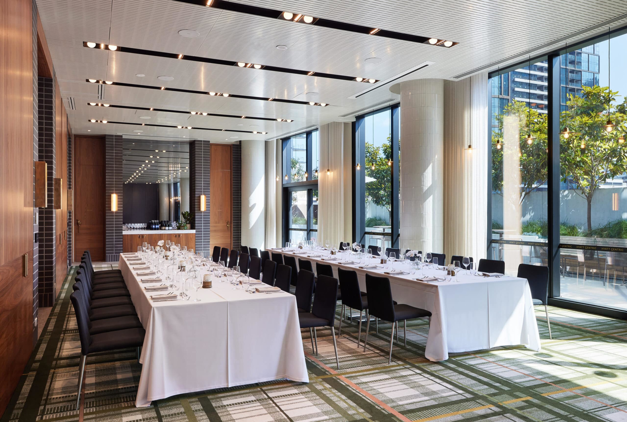 Modern indoor wedding reception room with long banquet tables and large windows overlooking a city terrace.