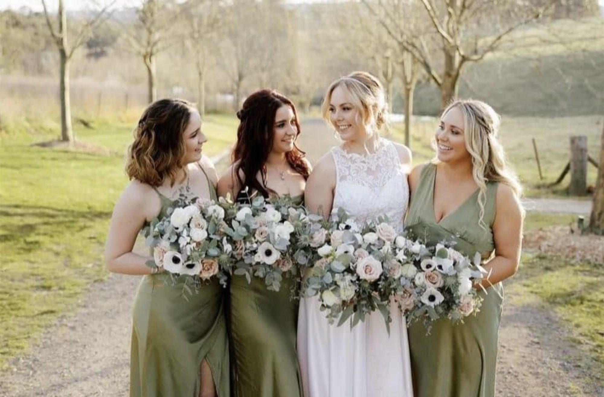 Bride and bridesmaids in green dresses holding lush bouquets while smiling together on a sunlit outdoor path.