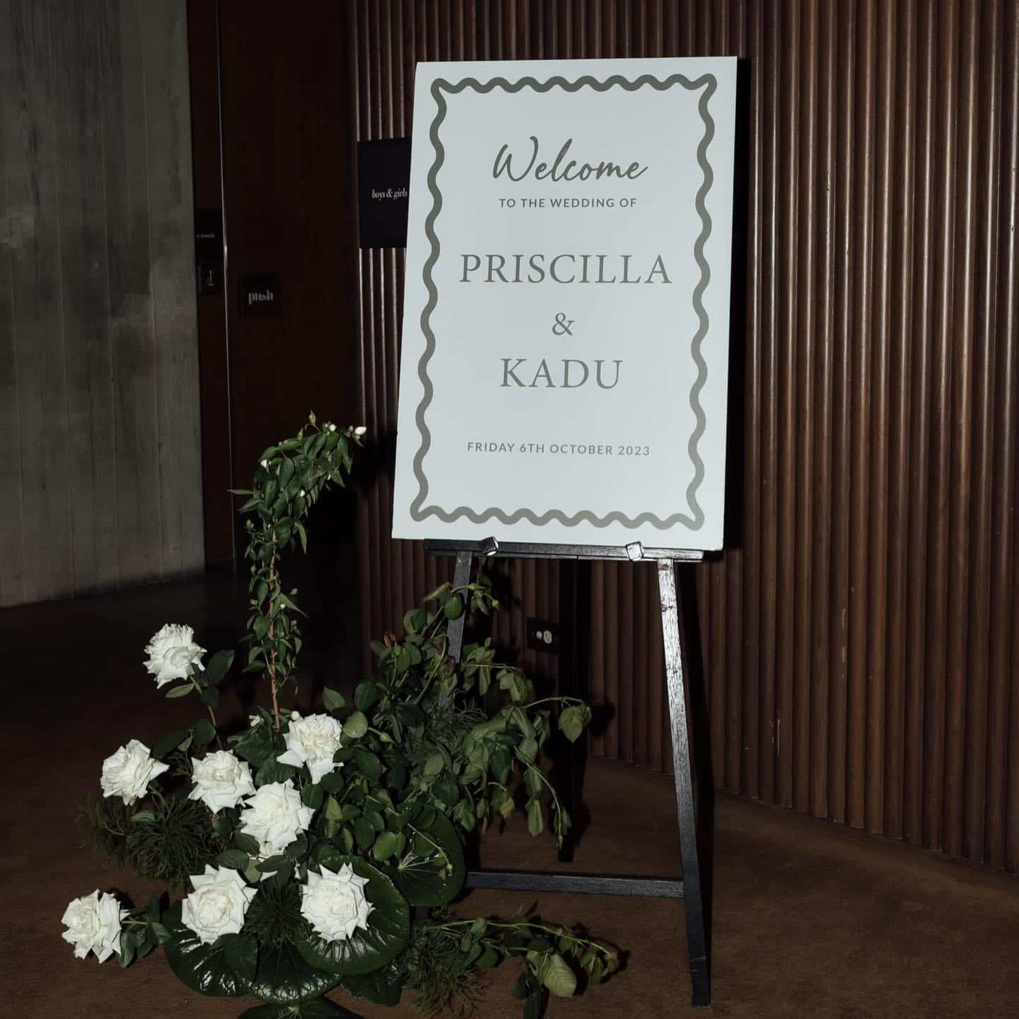 Modern wedding welcome sign for Priscilla and Kadu with white florals and greenery at the venue entrance.