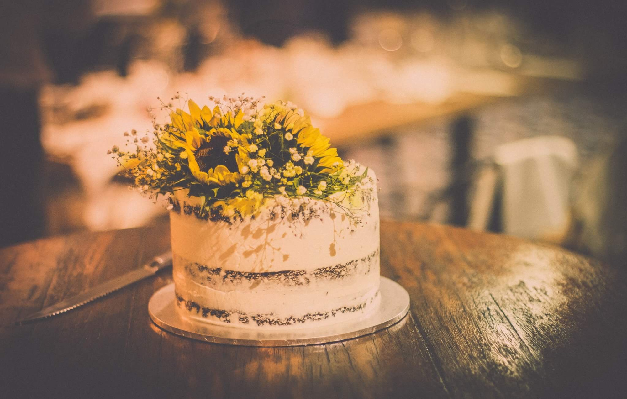 Rustic naked wedding cake topped with sunflowers and baby’s breath on a wooden table.