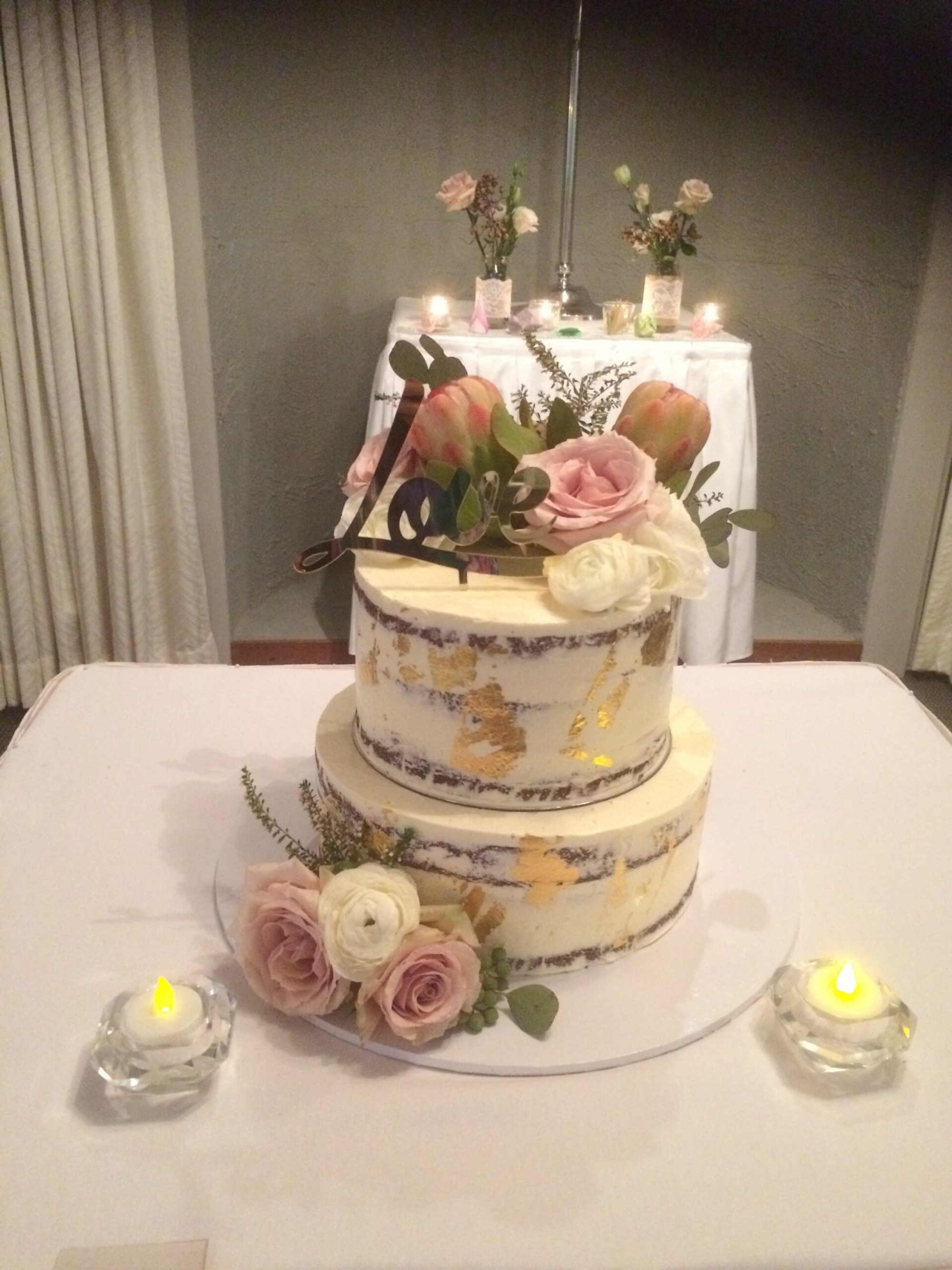 Two-tier semi-naked wedding cake with pink florals, gold leaf, and love topper surrounded by candles.
