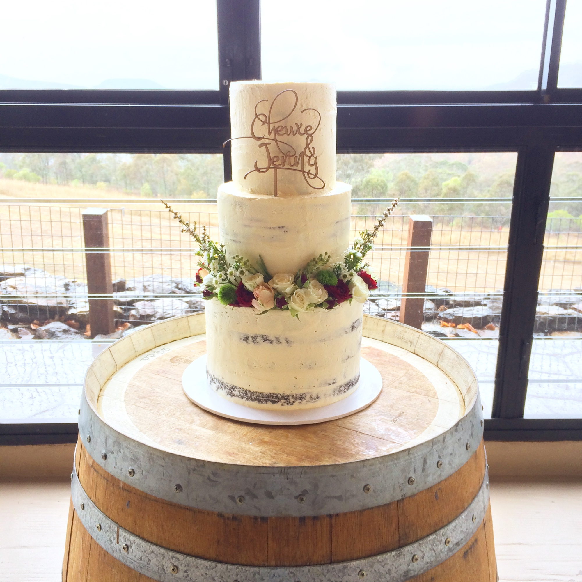 Three-tier semi-naked wedding cake with floral ring displayed on a wooden barrel in front of large windows.
