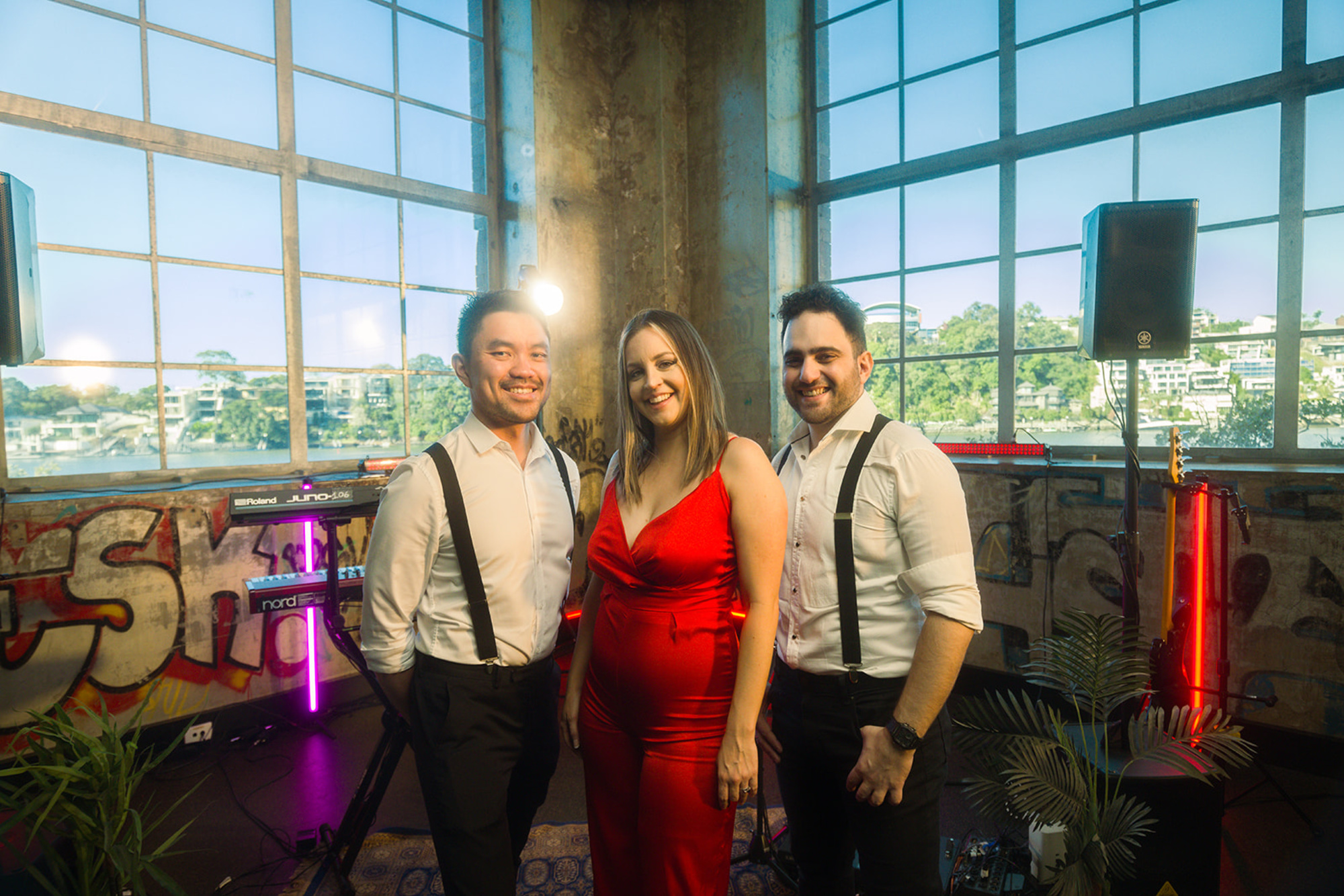 Three wedding band members pose in an industrial loft venue with large windows and musical equipment.