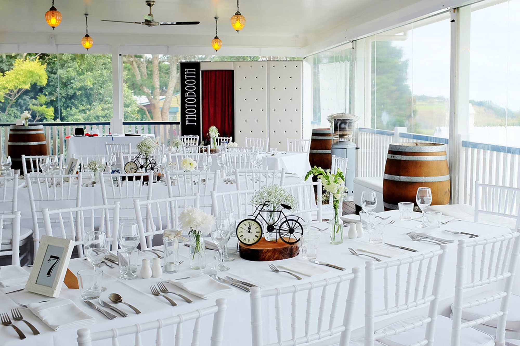Bright indoor wedding reception setup with white tables, chairs, and rustic barrel accents overlooking greenery.