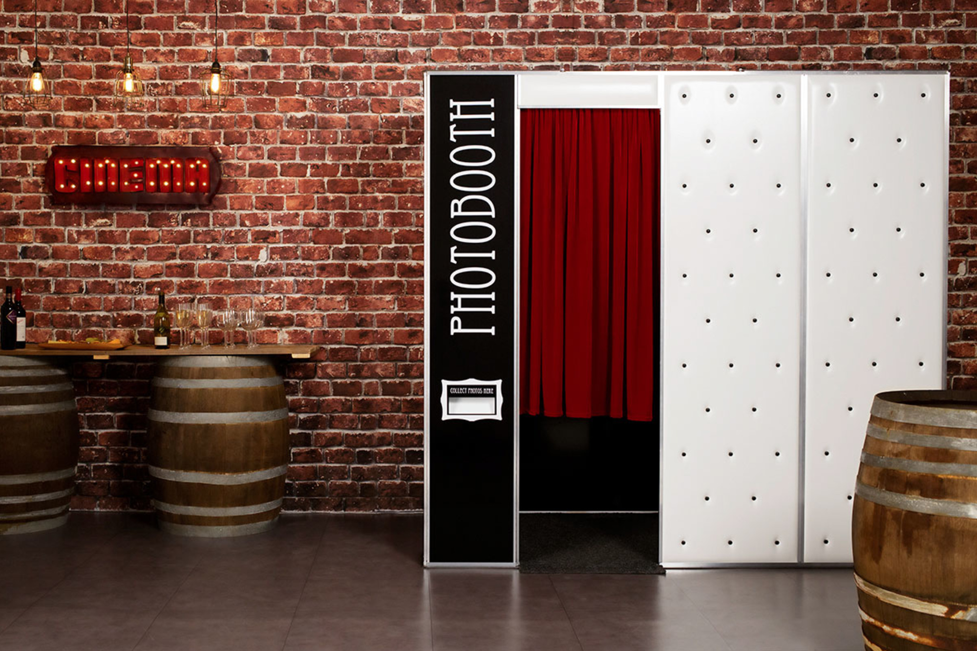 Rustic wedding photobooth setup with red curtain, brick wall backdrop and barrel bar with drinks and glassware.