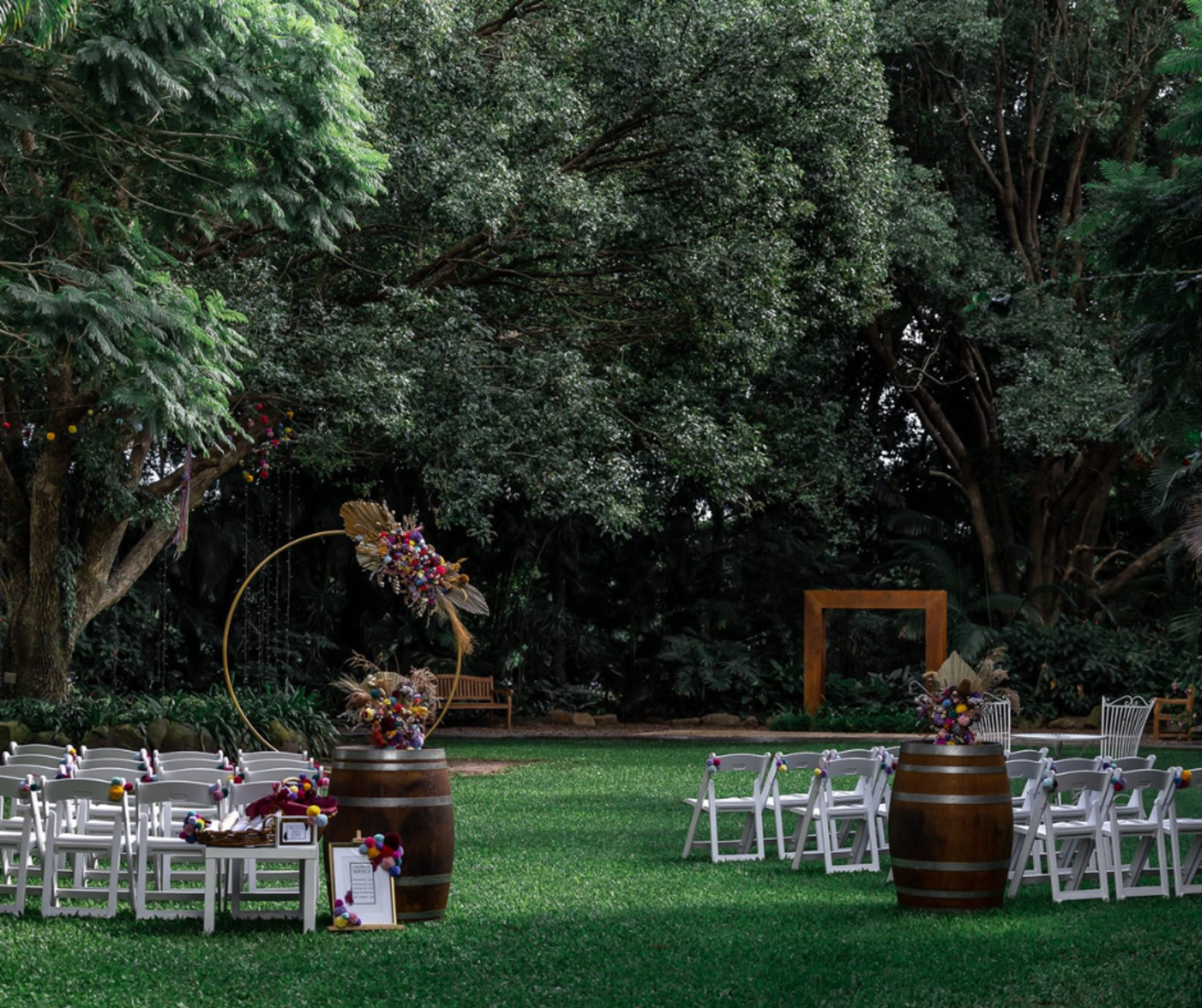 Outdoor wedding ceremony setup with white chairs, floral barrel decor, and wooden arch under large trees.