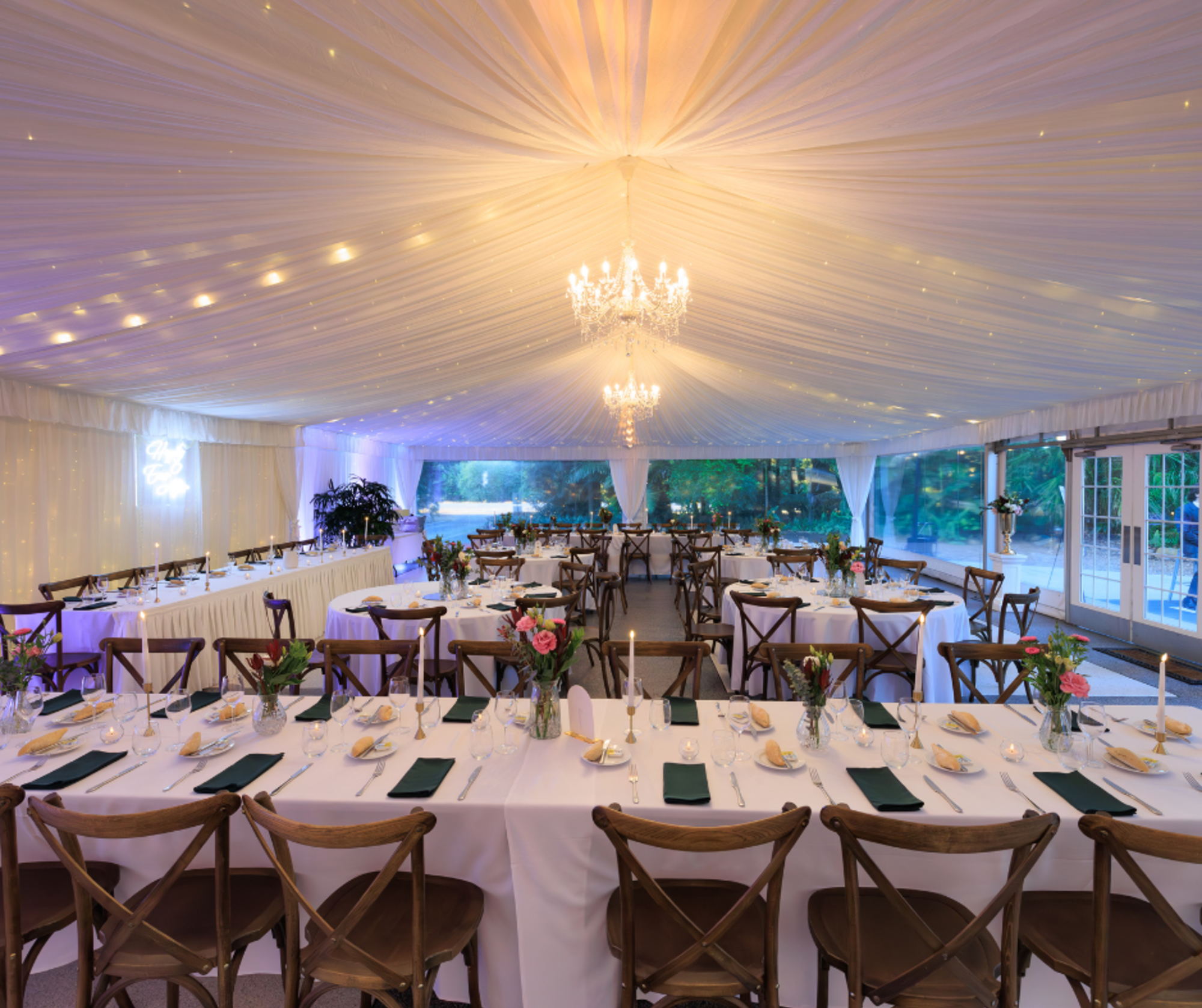 Elegant wedding reception setup in a draped tent with chandeliers, long banquet tables, and romantic candlelight.