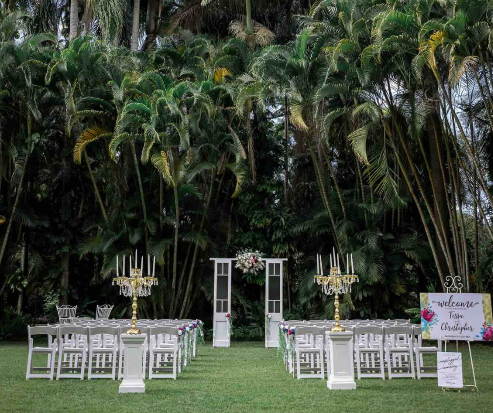 Outdoor wedding ceremony setup with white chairs, gold candelabras, and a floral-adorned doorway under tall palm trees.