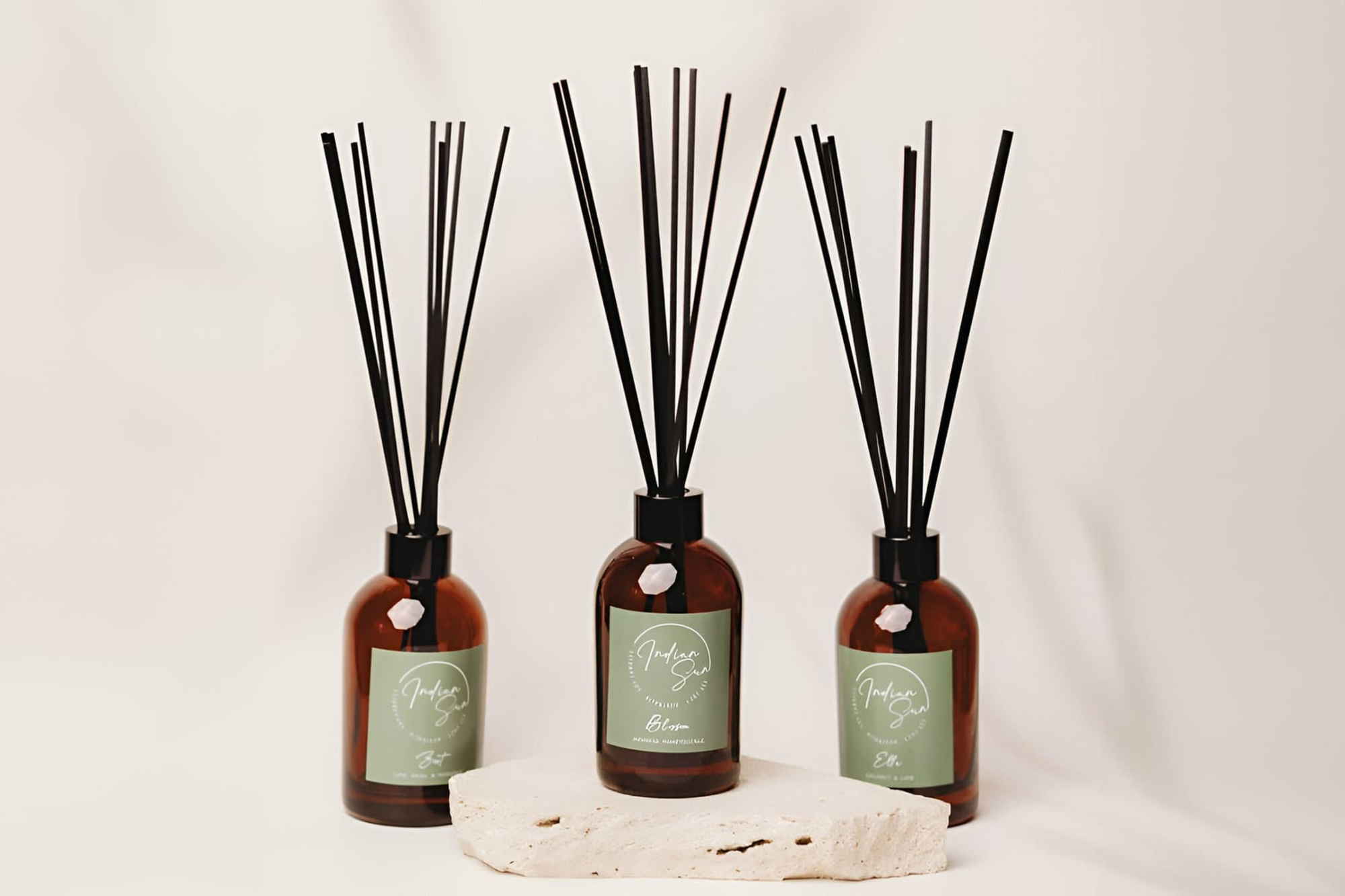 Three amber reed diffusers with green labels arranged on a neutral stone block against a soft beige backdrop.