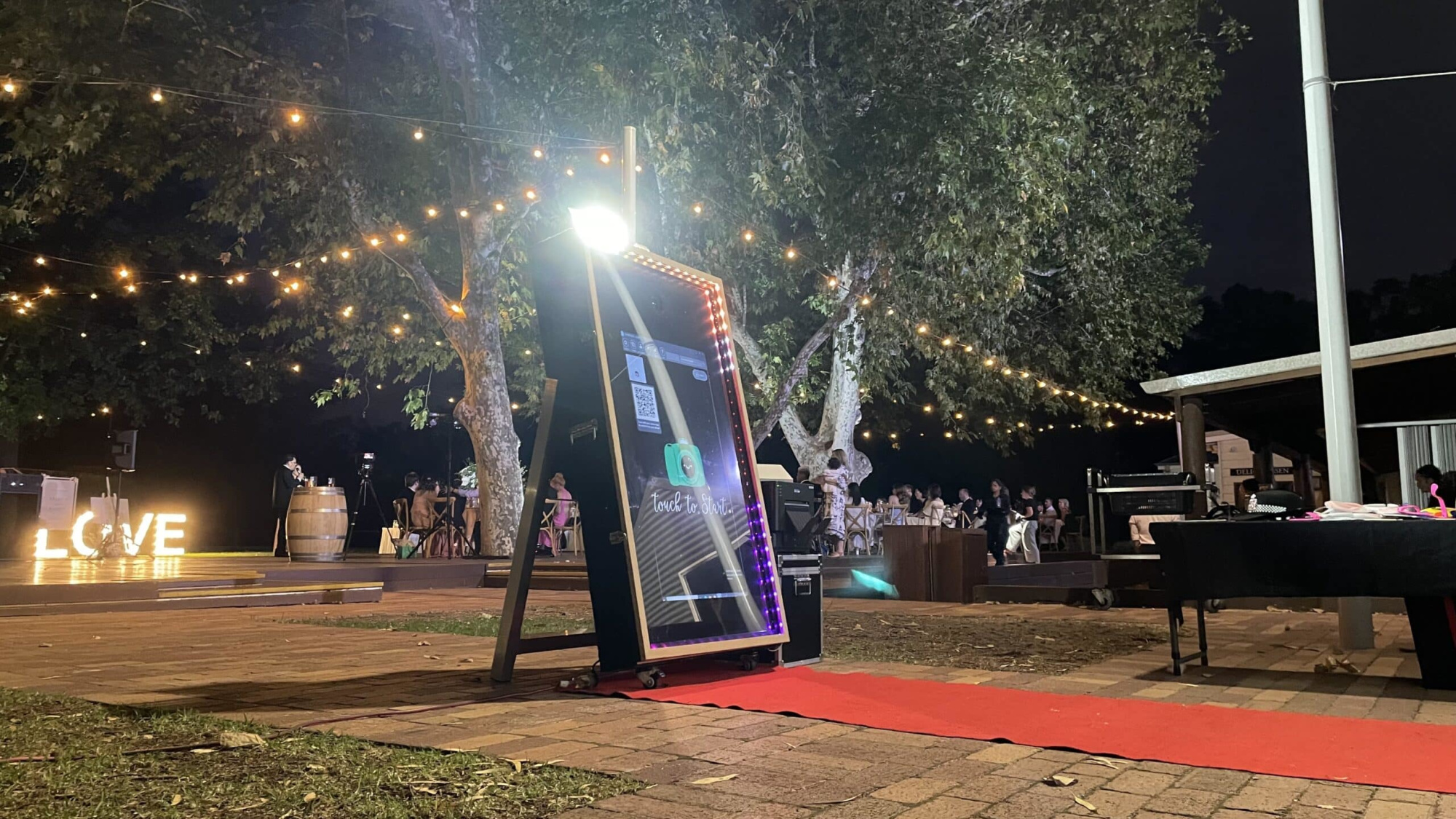 Outdoor evening wedding reception with a mirror photo booth, red carpet, and string lights under large trees.