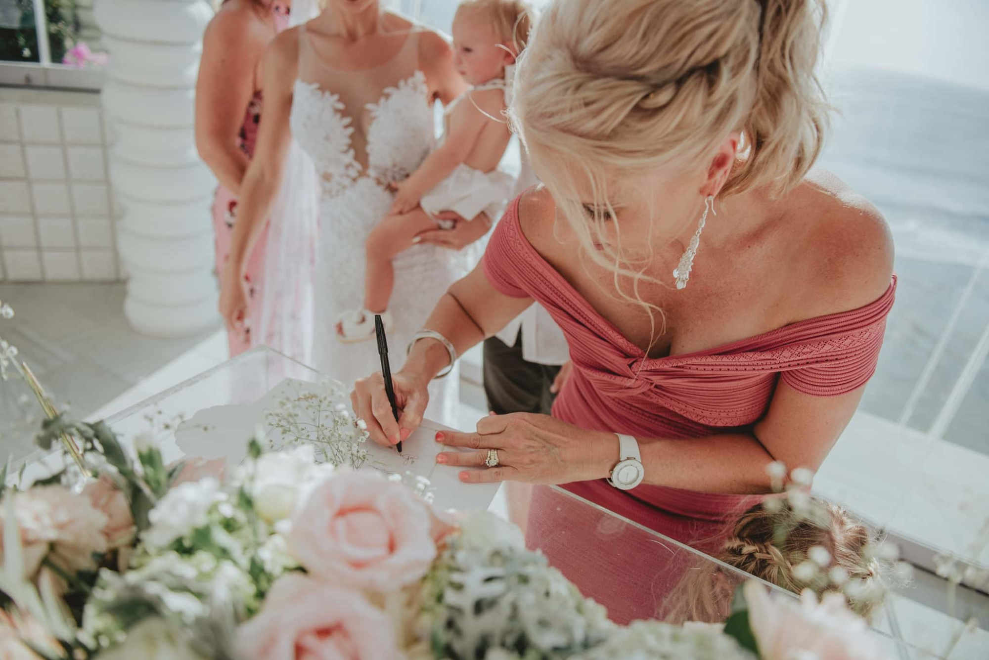 Woman in a rose dress signs a wedding document beside pastel flowers with the bride and child in the background.