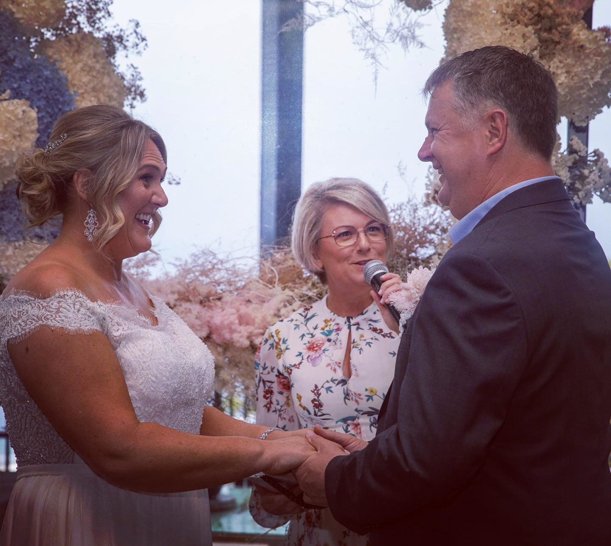 A wedding celebrant leads a couple exchanging vows in front of a soft floral backdrop.