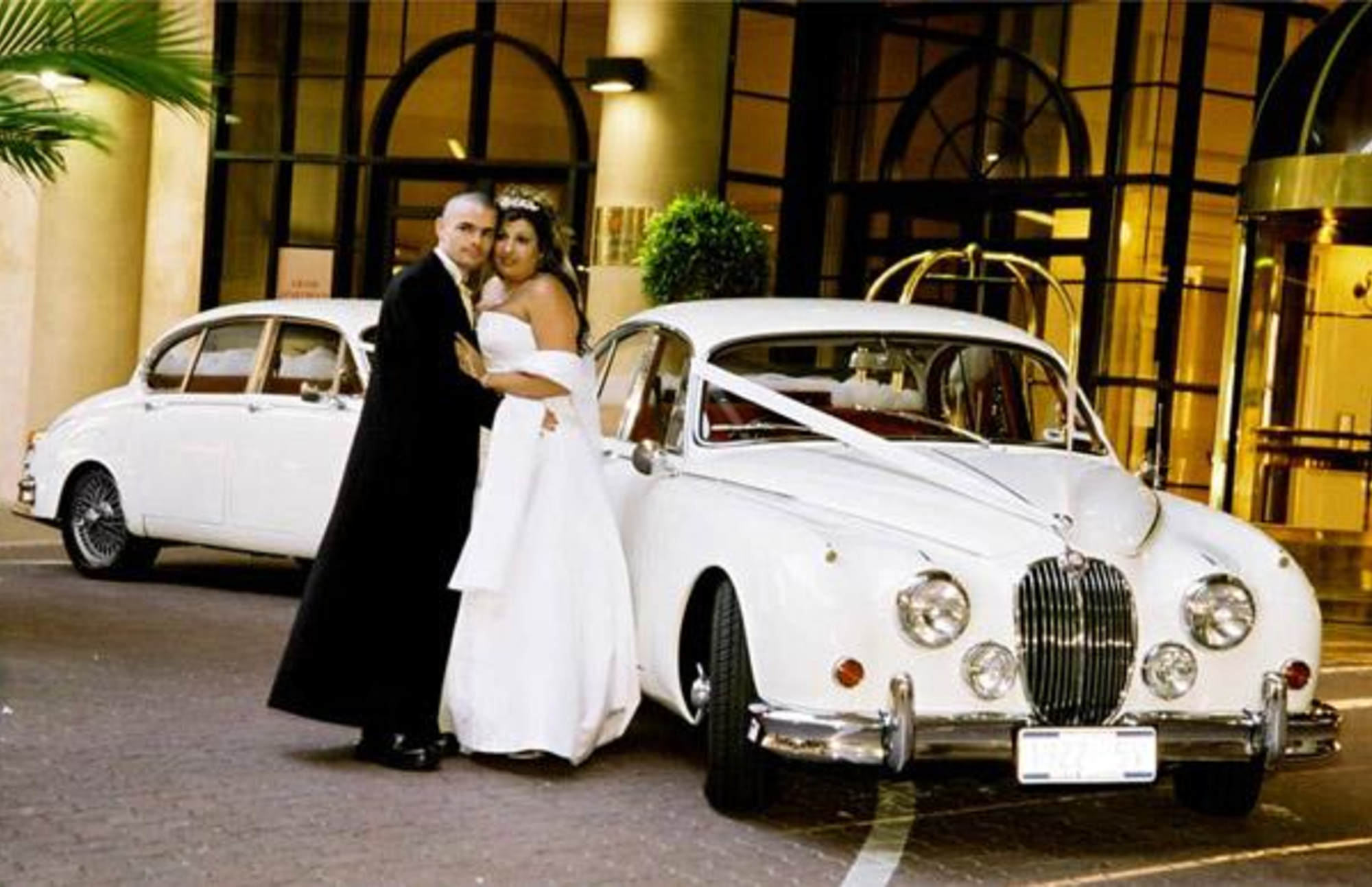 Bride and groom pose beside a white classic wedding car outside an elegant hotel entrance.