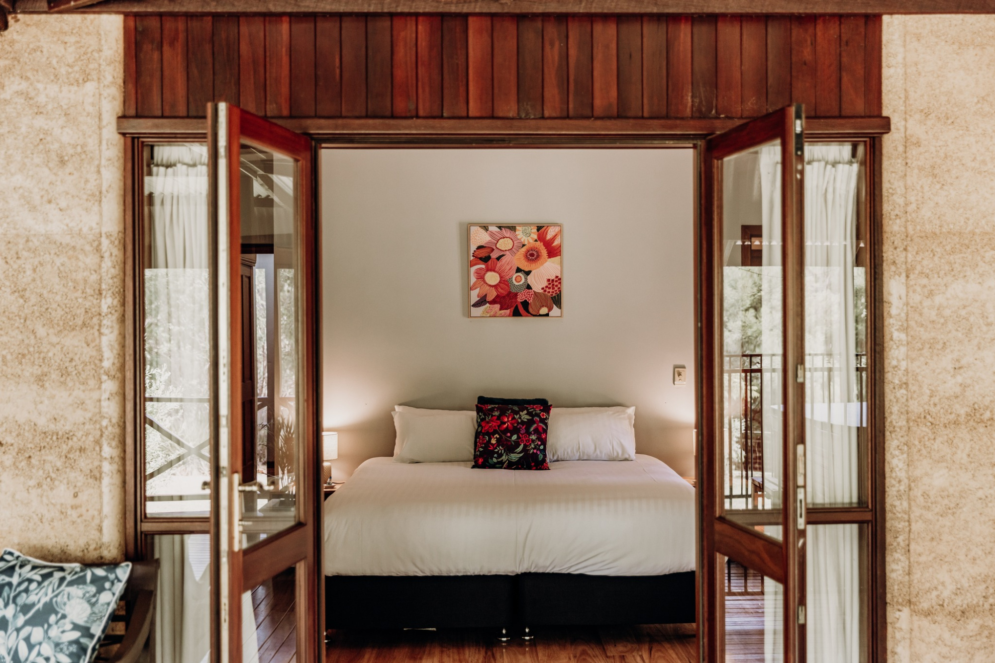 Cozy rustic bedroom with a white bed, floral artwork, and open wooden doors leading to a balcony.